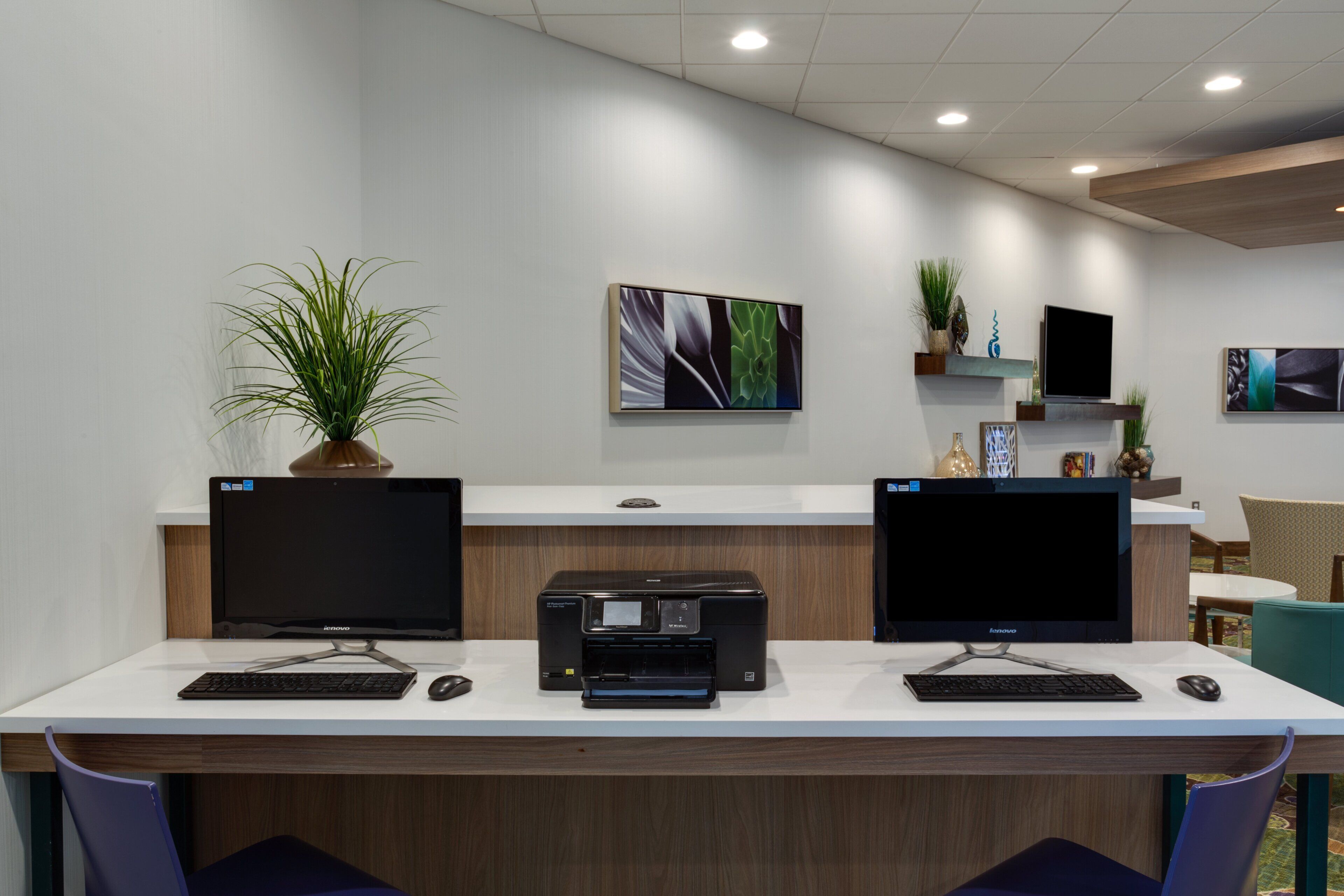 A modern office space with two desks, each with a computer monitor, keyboard, and mouse. There are two printers on the desks, and the walls are adorned with framed artwork.