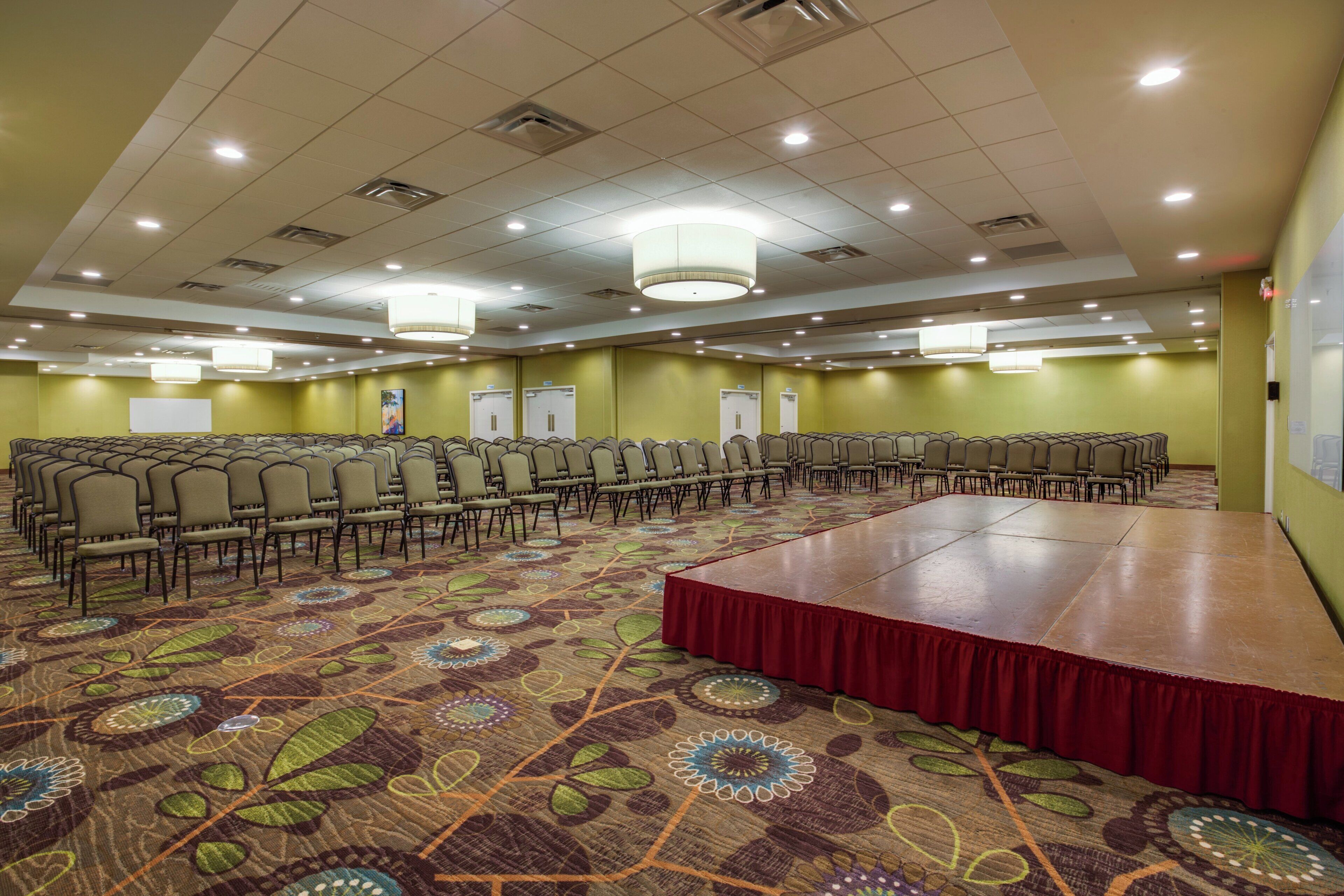 A conference room with rows of chairs, a stage with a red carpet, and a patterned carpet.