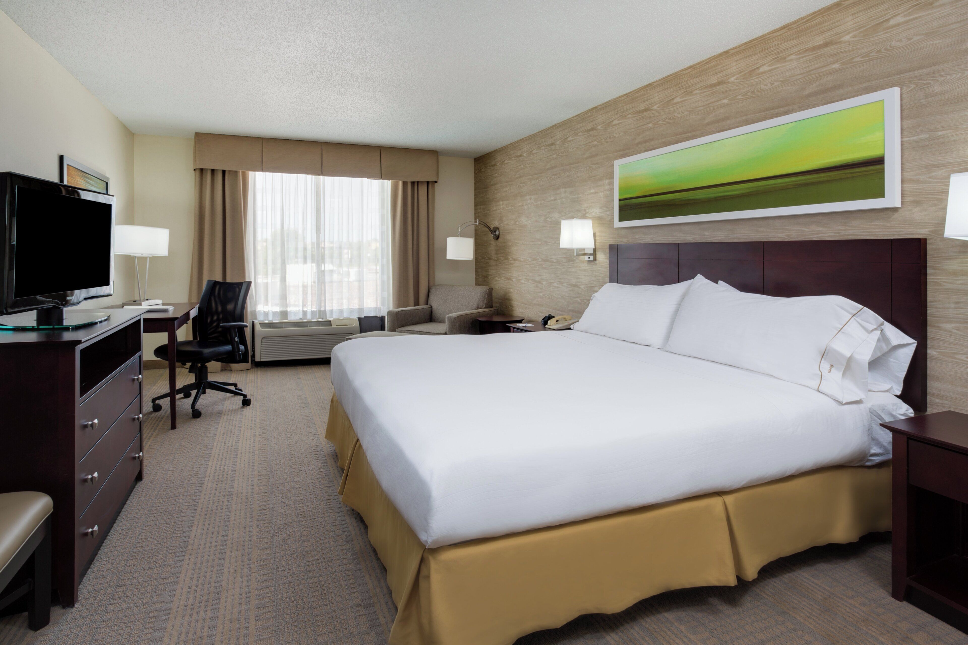 Habitación de hotel con una cama grande, un escritorio con televisor, una silla y una ventana con cortinas.