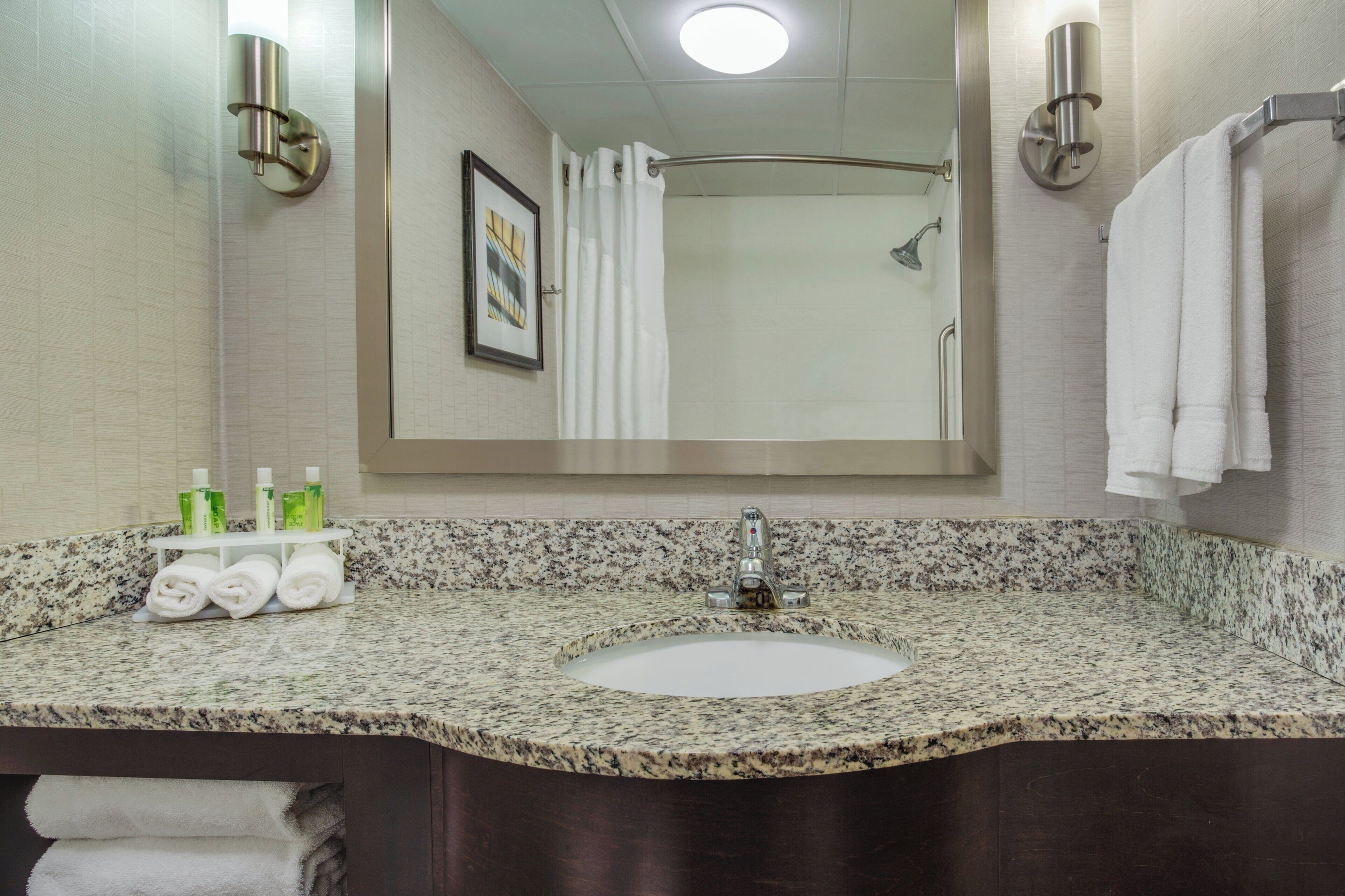 Une salle de bain avec un lavabo doté d’un plan de travail en granit, un miroir, un porte-serviettes et un tableau encadré au mur.