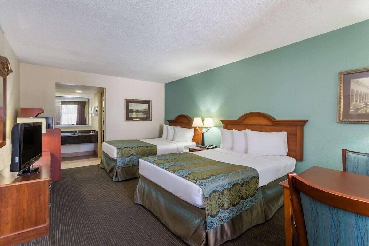 Habitación de hotel con dos camas, televisión, escritorio y baño visible a través de una puerta abierta.