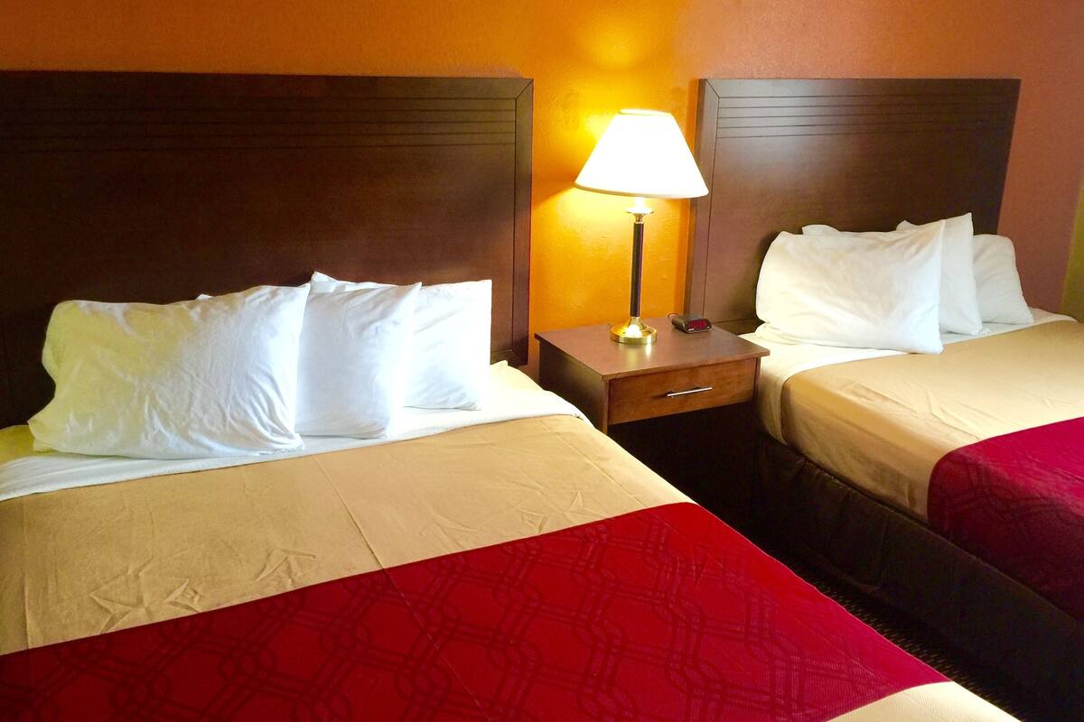 Quarto de hotel com duas camas, cada uma com lençóis brancos e cobre vermelhos. Há uma mesinha de cabeceira com abajur entre as camas.