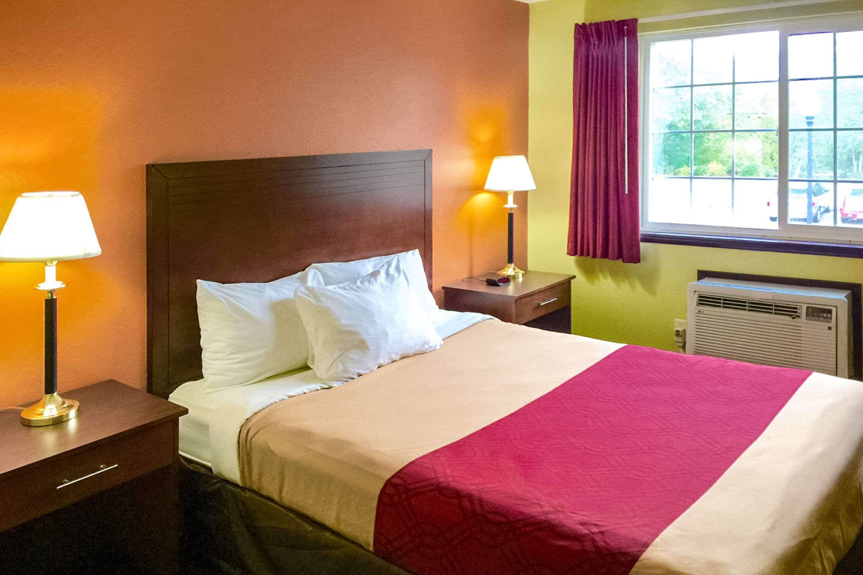 A hotel room with a bed, two bedside lamps, a window with curtains, and an air conditioning unit.