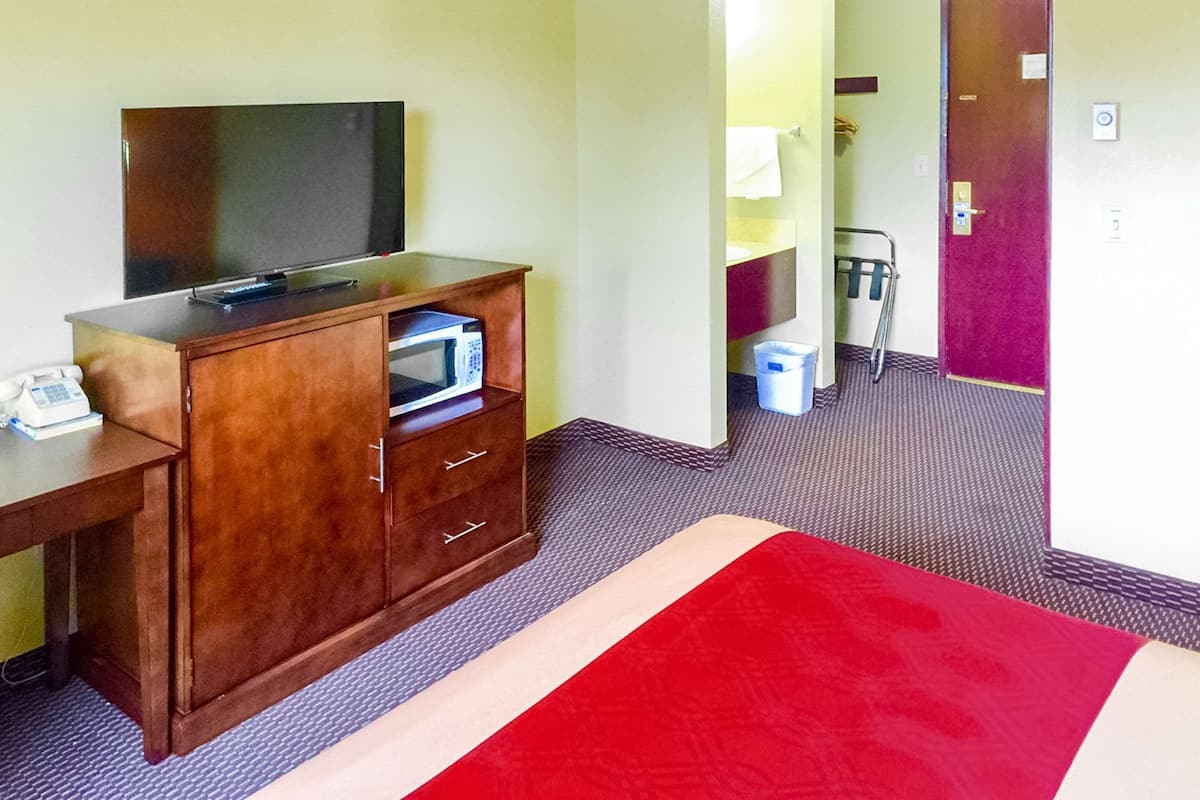 Quarto de hotel com escrivaninha de madeira, televisão de tela plana, micro-ondas e tapete vermelho.