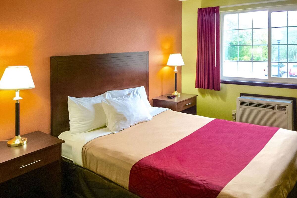 Quarto de hotel com cama, duas luminárias de cabeceira, mesa de cabeceira, janela com cortinas e aparelho de ar condicionado.