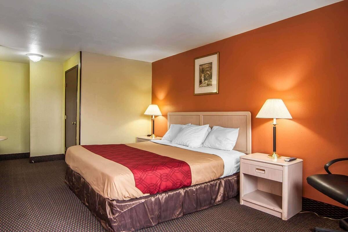 Quarto de hotel com cama, duas luminárias de cabeceira, mesa de cabeceira, uma cadeira e uma porta.