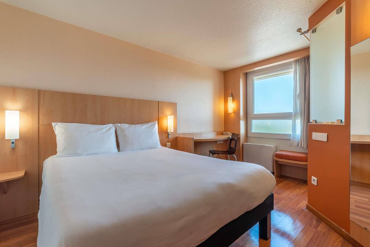A hotel room with a large bed, a desk, and a chair. There is a window with curtains and a view of the outside.