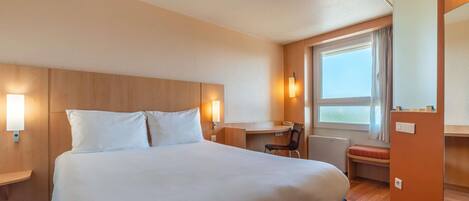 Habitación de hotel con una cama grande, un escritorio y una silla. Se ve una ventana con cortinas y vistas al exterior.