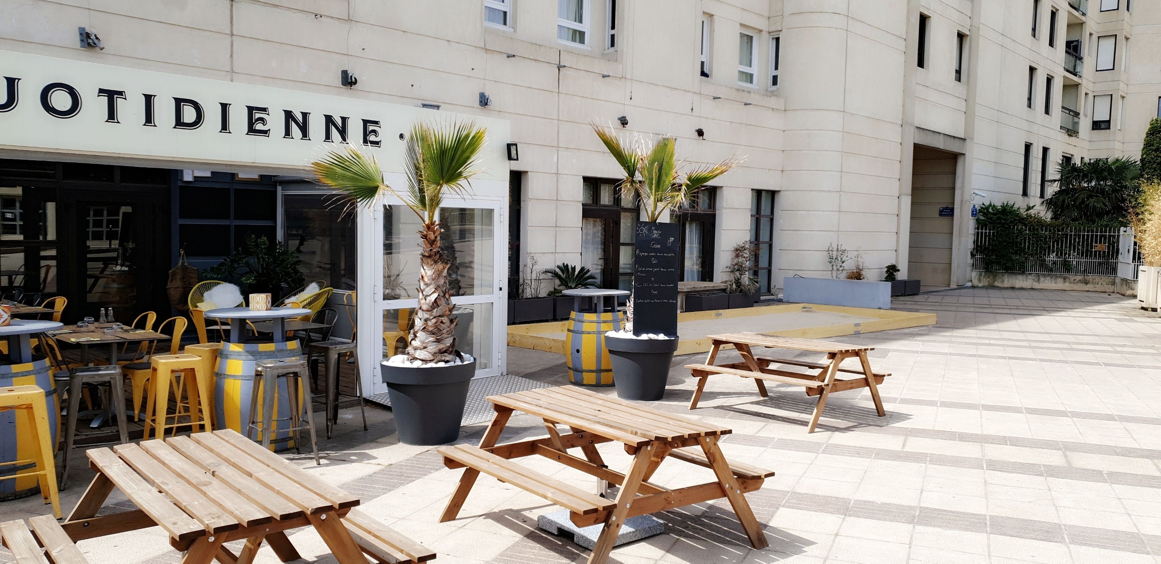 An outdoor seating area with wooden tables and chairs, potted plants, and a building with the sign "JOTIDIENNE".