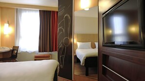 Ein Hotelzimmer mit zwei Betten, einem Schreibtisch, einem Sessel, einem großen Spiegel und einem Fernseher.