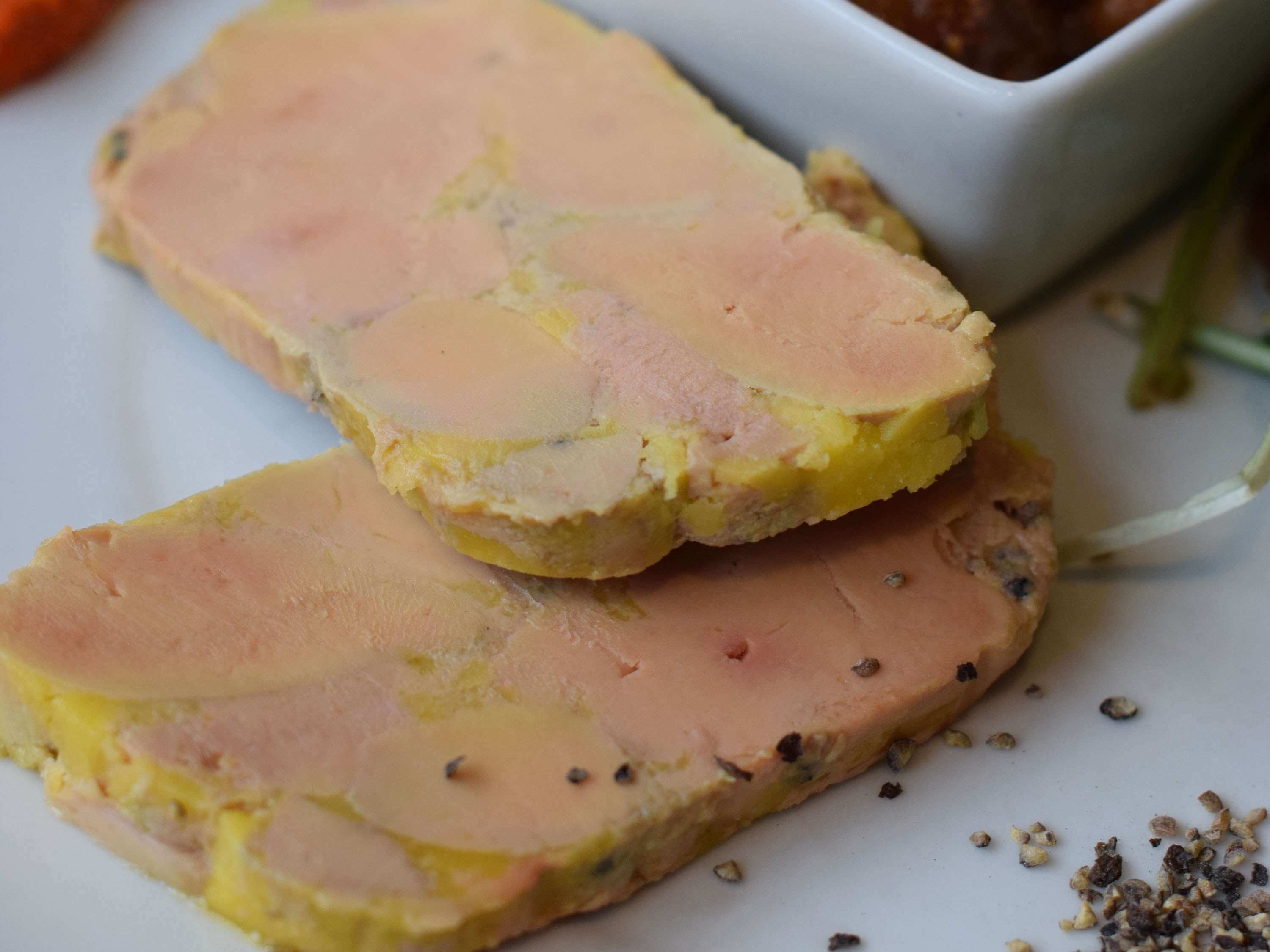 A plate with slices of a layered meat dish, possibly a terrine, with a creamy yellow filling and a pink outer layer, accompanied by a small bowl of sauce.