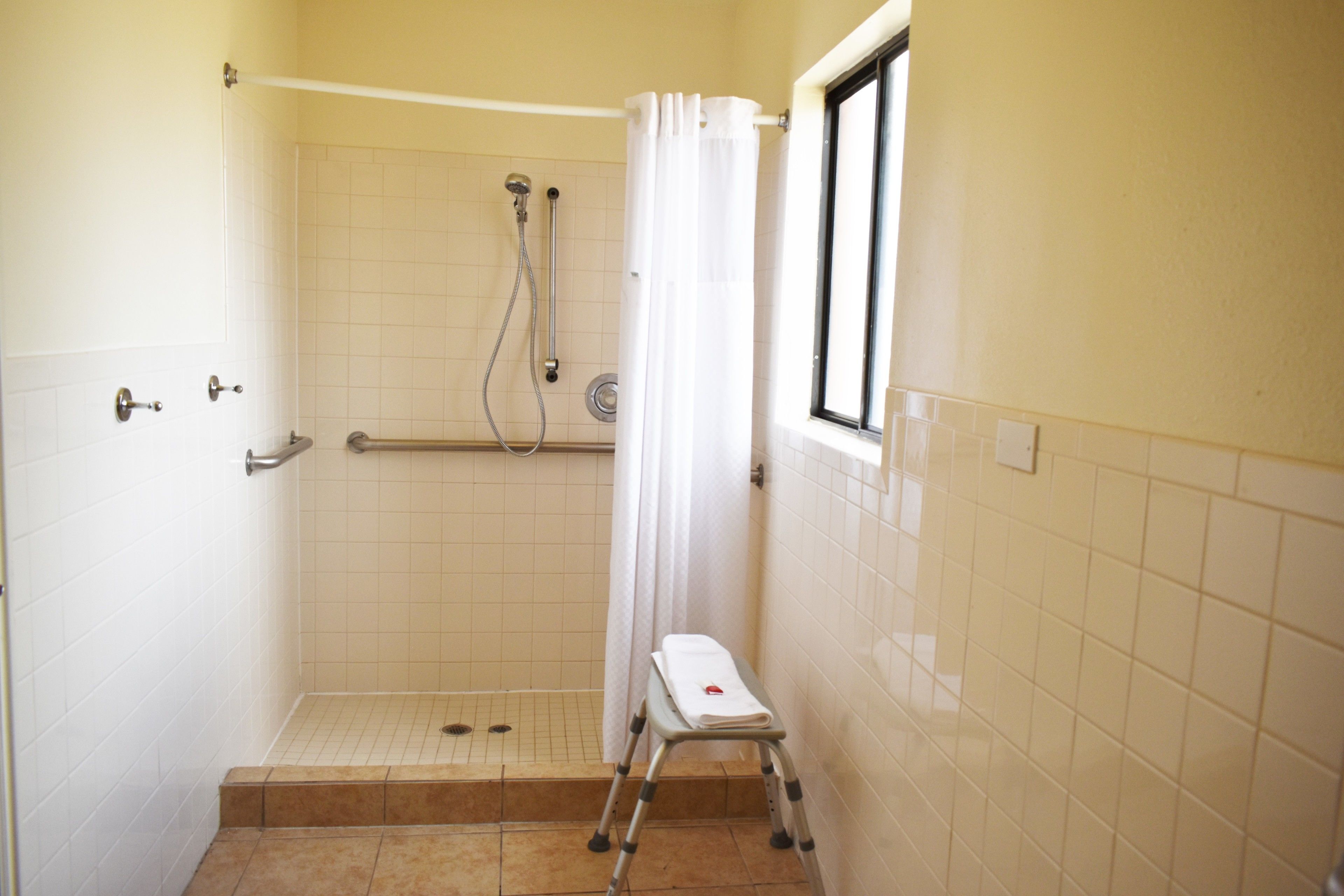 A small, tiled shower area with a window, a showerhead, a handrail, and a step stool.