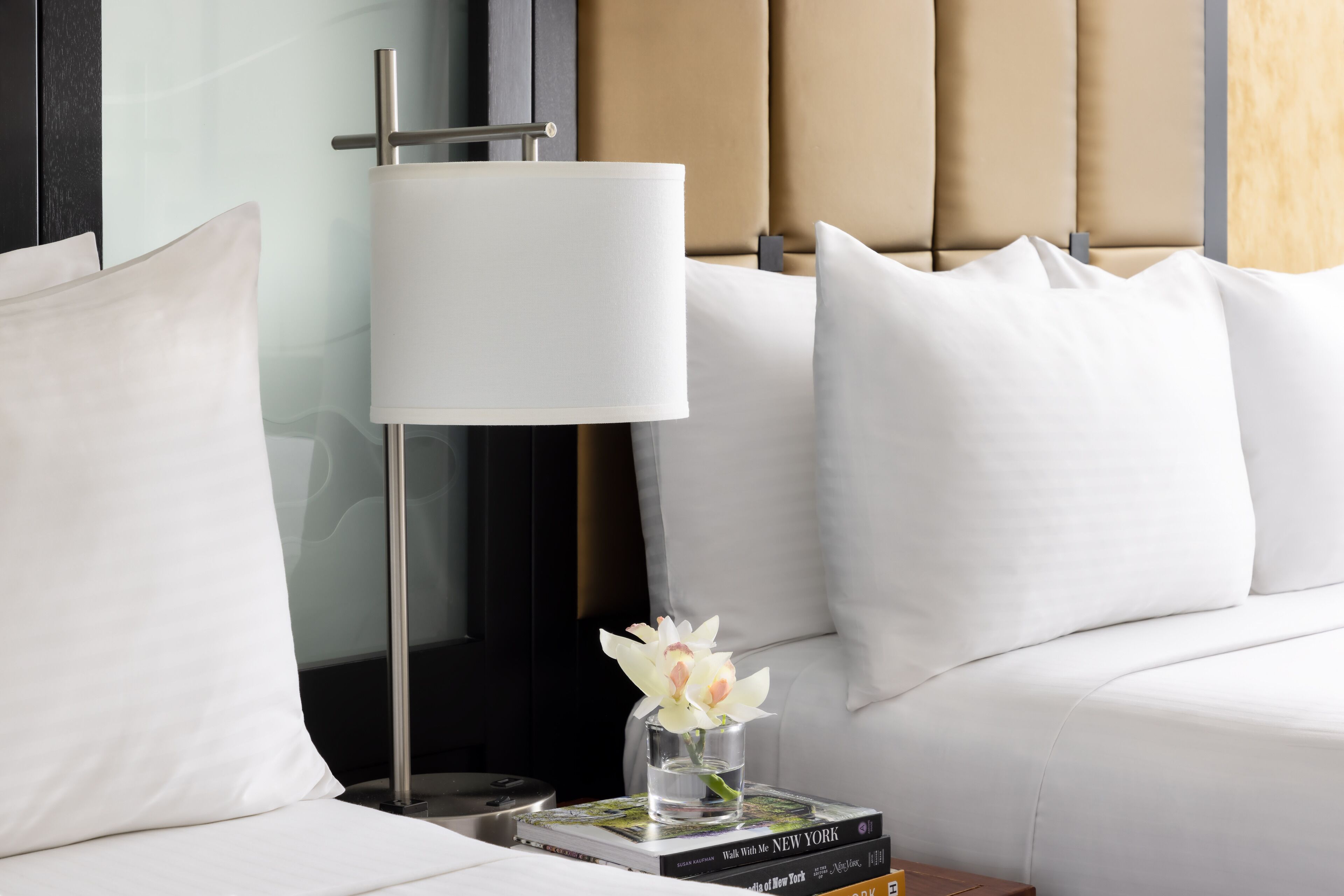 Una cama bien tendida con sábanas blancas, una lámpara de noche, un florero con una sola flor y una pila de libros en la mesita de noche.