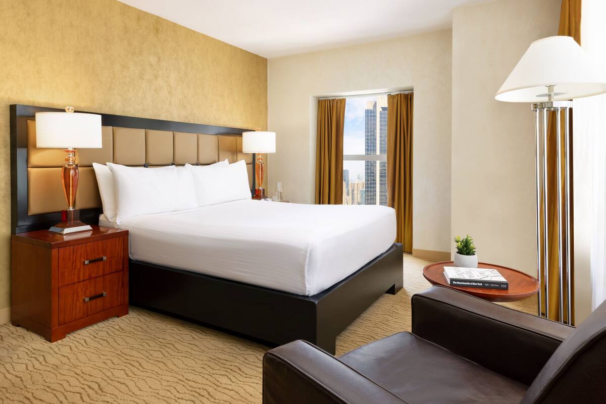 A hotel room with a large bed, two bedside lamps, a small table with a book and a plant, and a view of the city through a window.