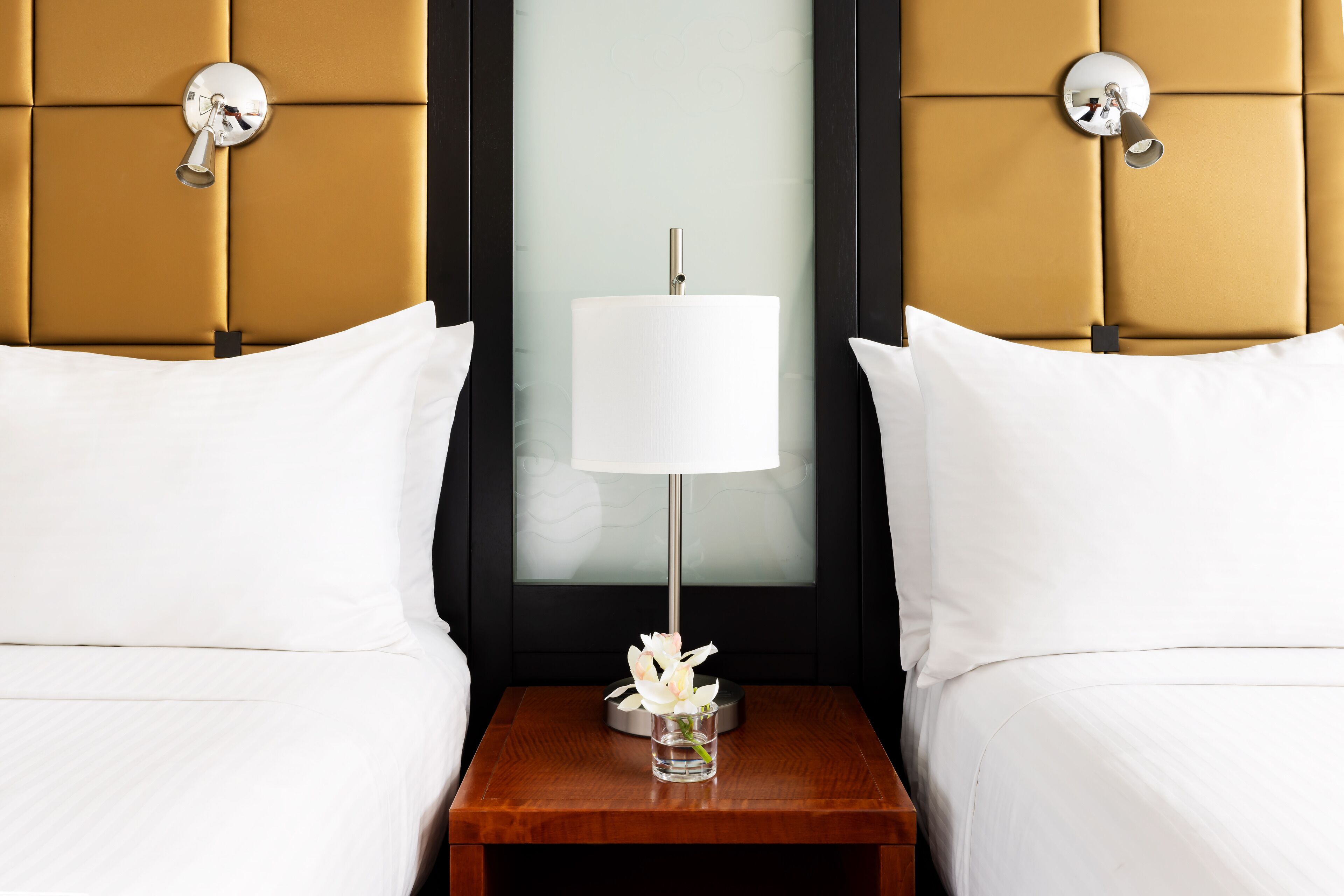 Una habitación de hotel con cabecera capitonada, una mesita de noche con lámpara y ropa de cama blanca.