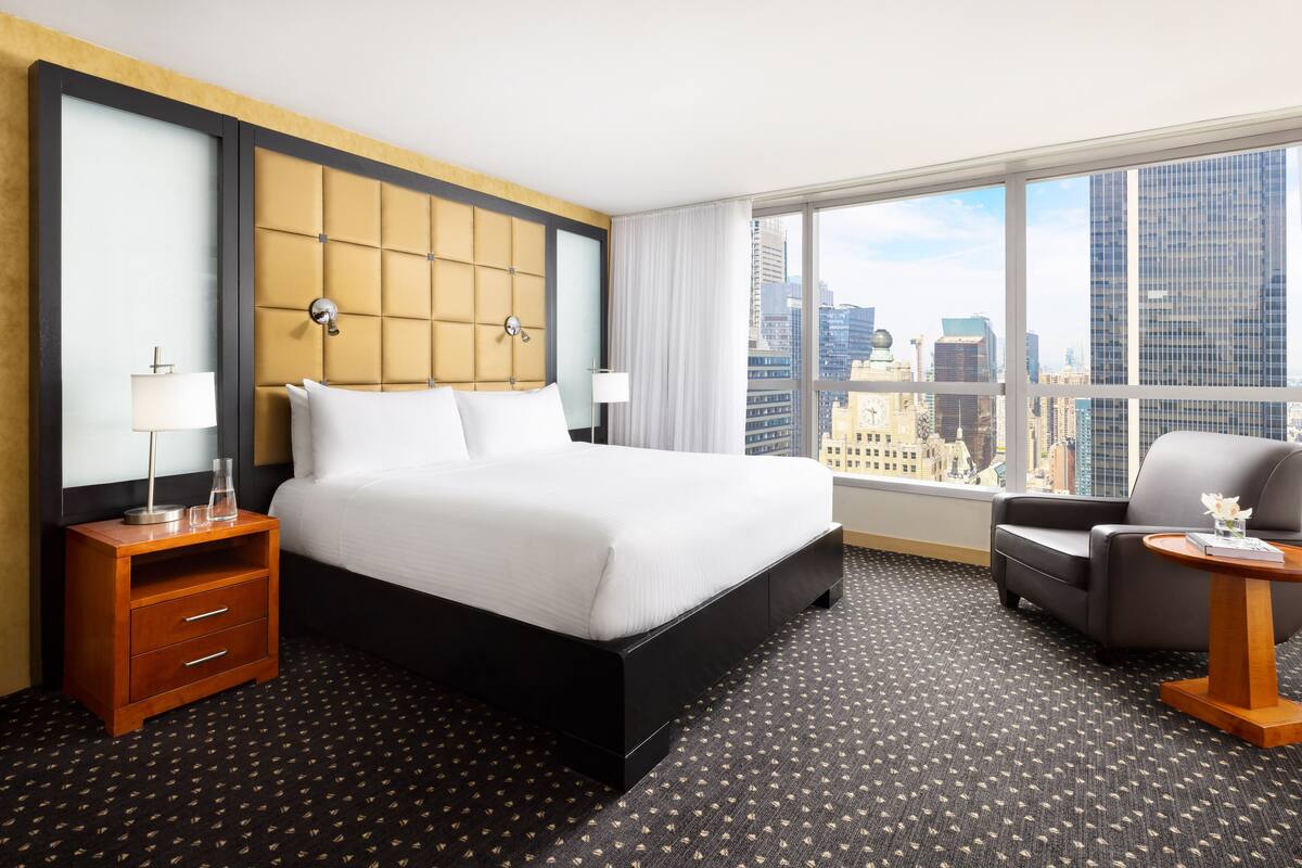 A hotel room with a large bed, a nightstand with a lamp, a chair, and a view of the city through large windows.