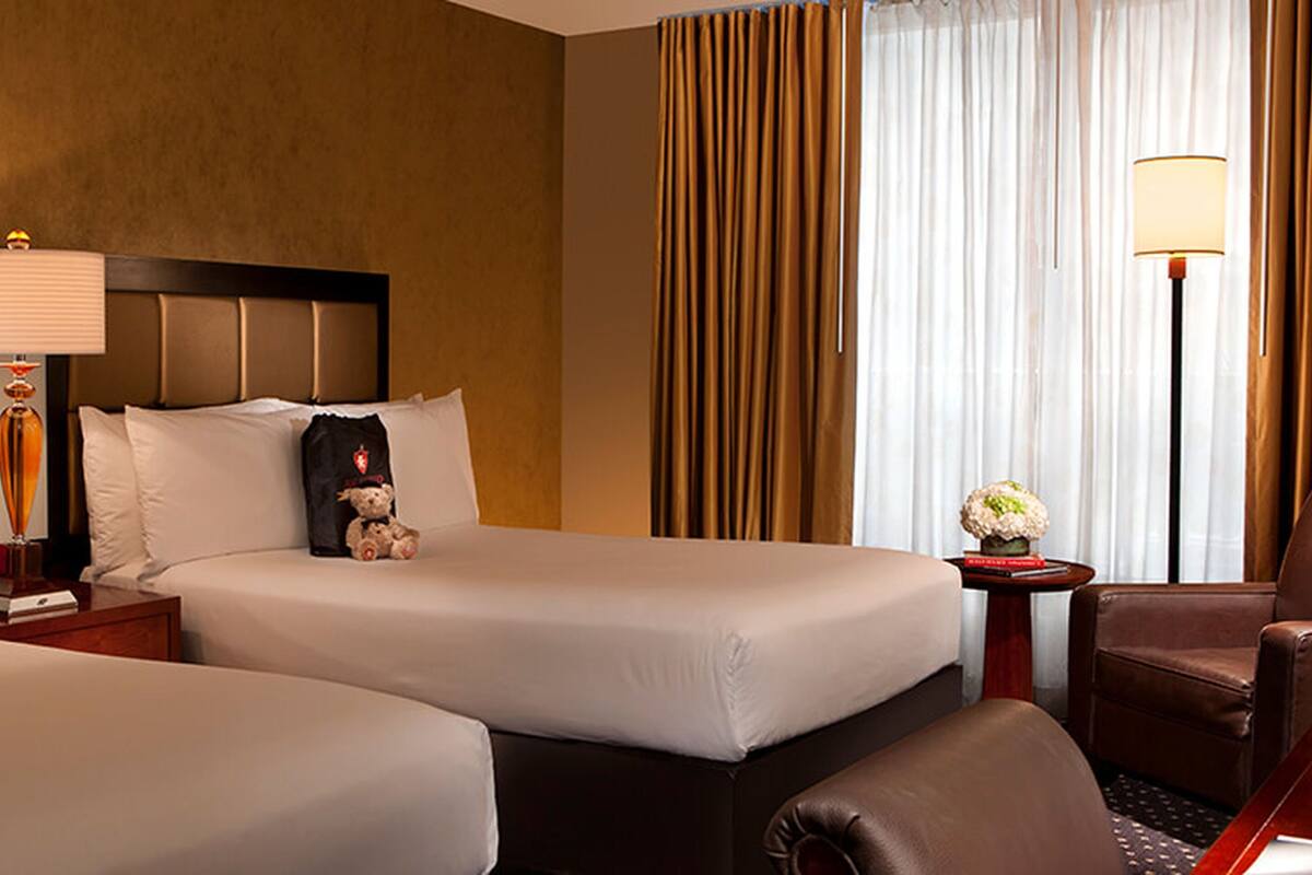 A hotel room with two beds, a nightstand with a lamp, a chair, and a desk with a lamp.