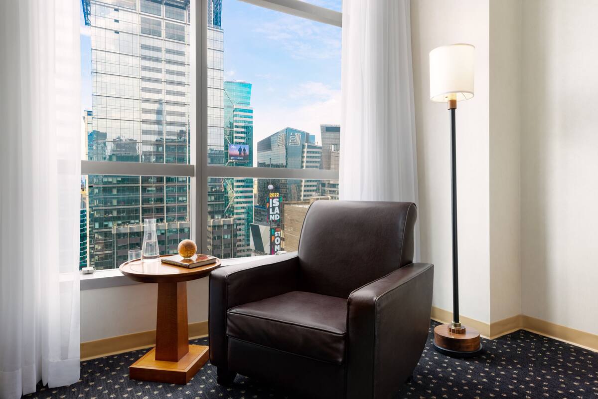 A hotel room with a large window offering a city view, a leather armchair, a small wooden side table, and a floor lamp.