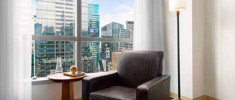 A hotel room with a large window offering a city view, a leather armchair, a small wooden side table, and a floor lamp.