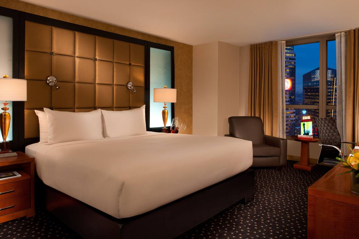 A hotel room with a large bed, two bedside lamps, a chair, a desk, and a view of the cityscape.