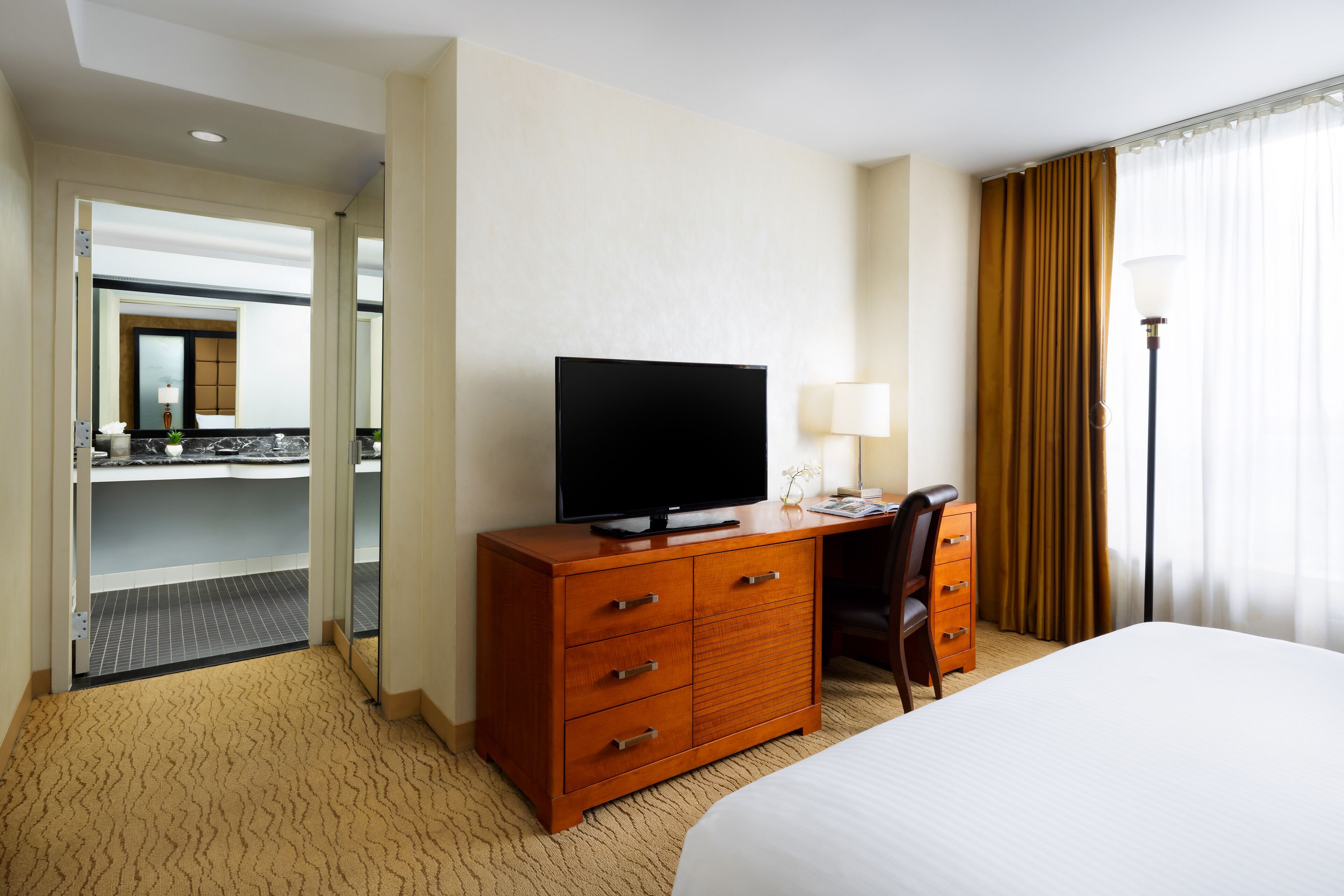 A hotel room with a bed, a wooden dresser with a flat-screen TV, a desk with a lamp, and a view of a balcony.