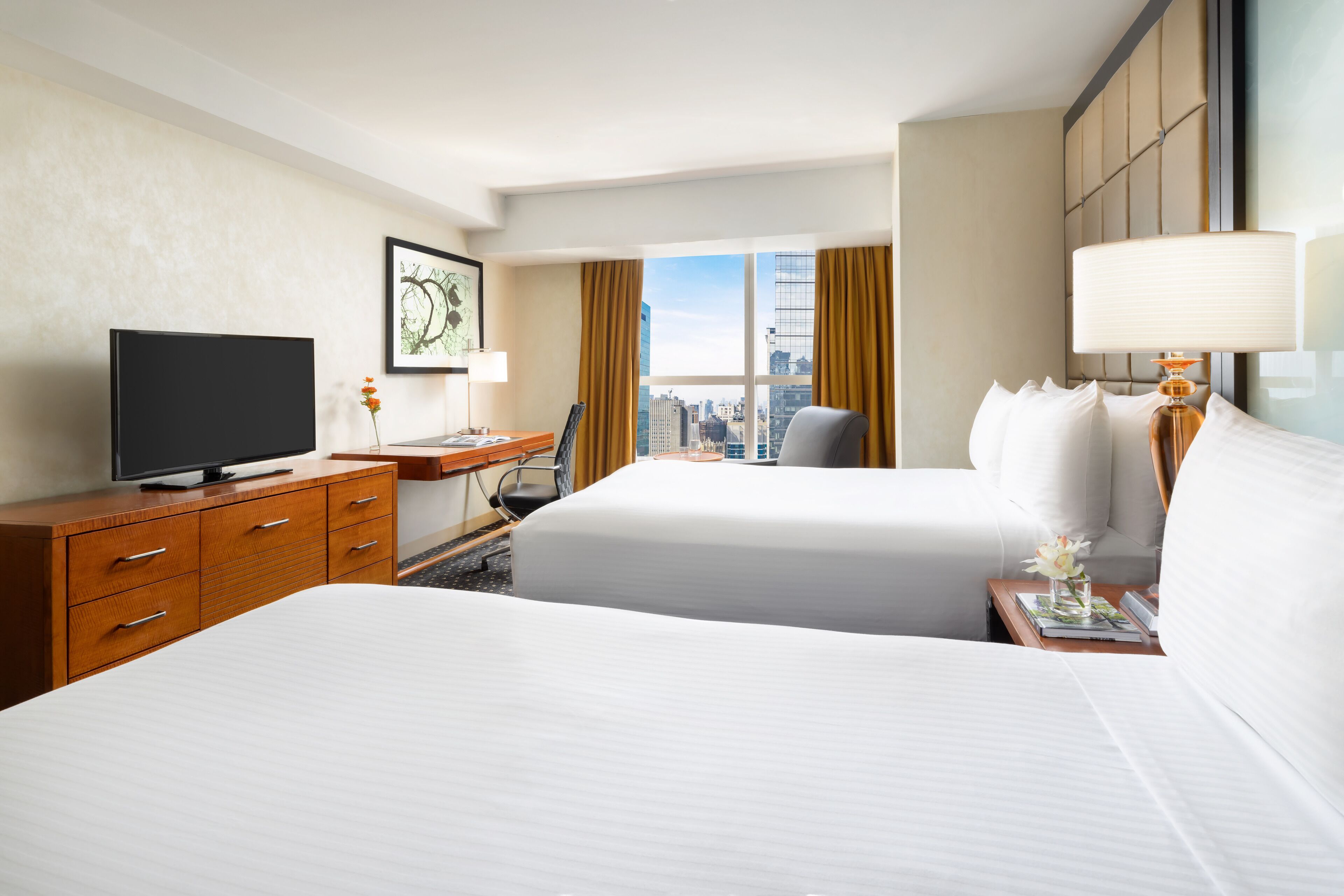 Ein Hotelzimmer mit zwei Betten, einem Schreibtisch, einem Fernseher und einem Fenster mit Blick auf die Stadt.