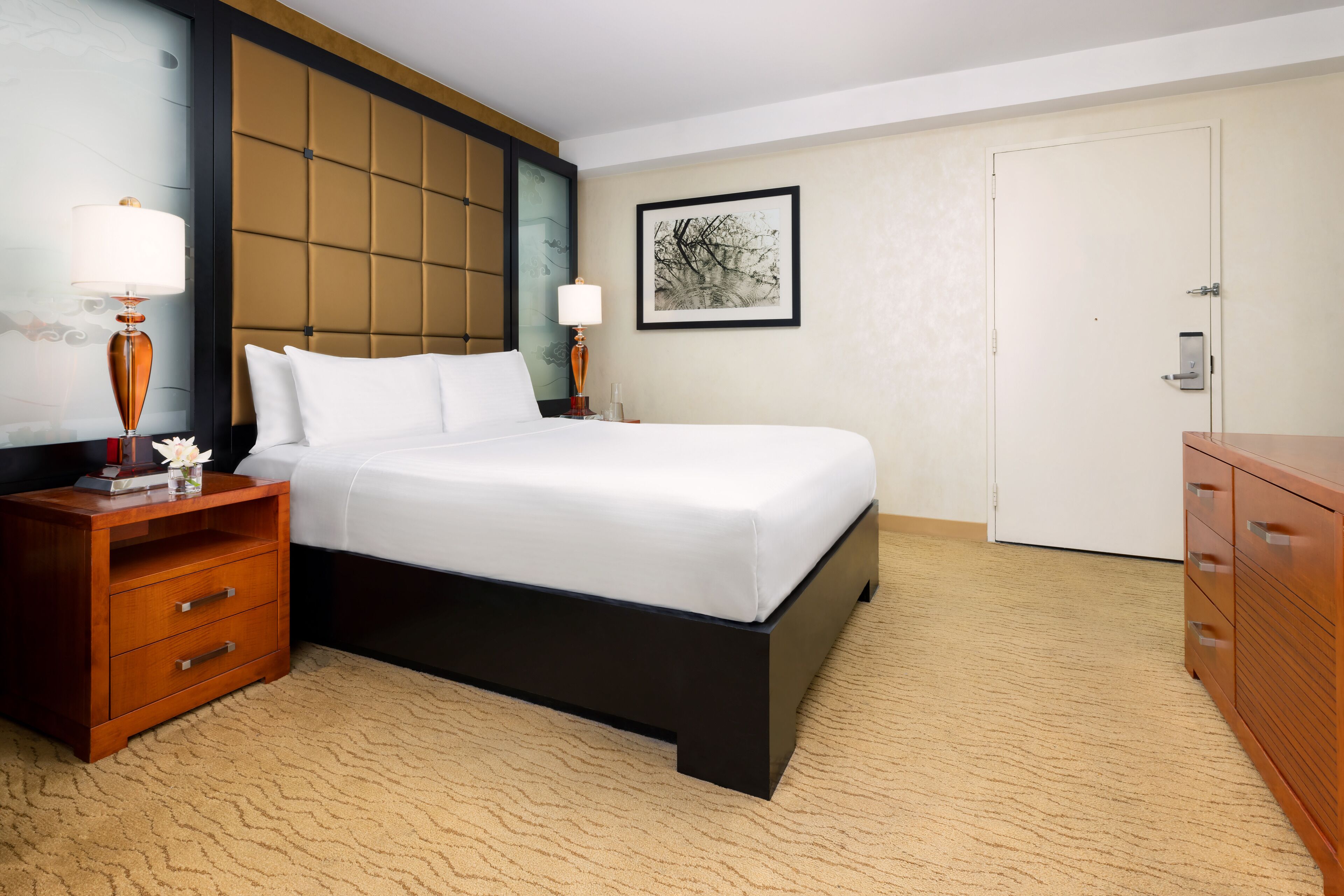 Une chambre d’hôtel avec un grand lit, une table de chevet avec une lampe, une commode et un tableau encadré au mur.