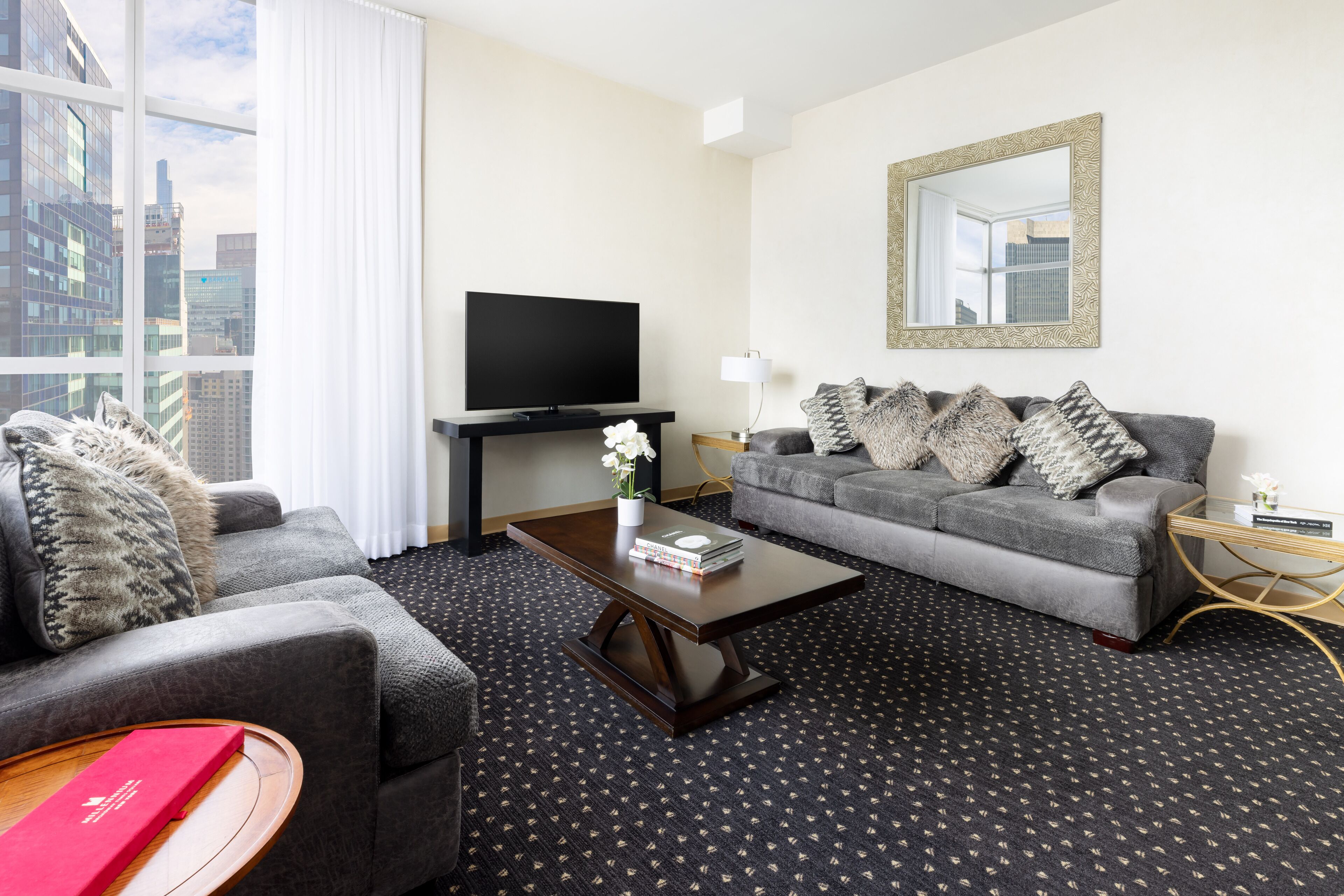A modern hotel room with a grey sofa, a smaller armchair, a wooden coffee table, a flat-screen TV, a large mirror, and a city view through the window.