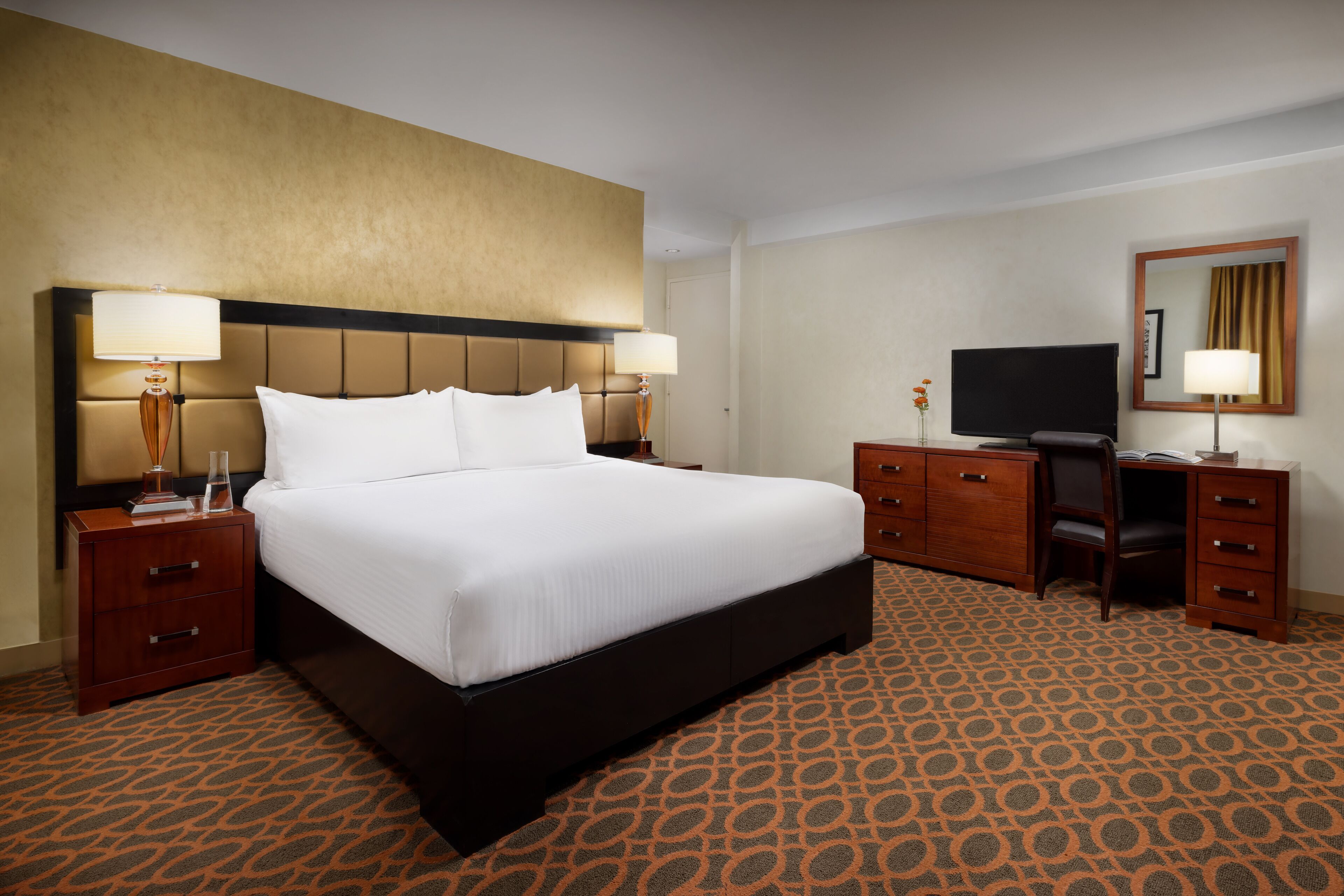 Quarto de hotel com uma cama grande, duas mesas de cabeceira com luminárias, uma escrivaninha com cadeira, televisão e um espelho na parede.