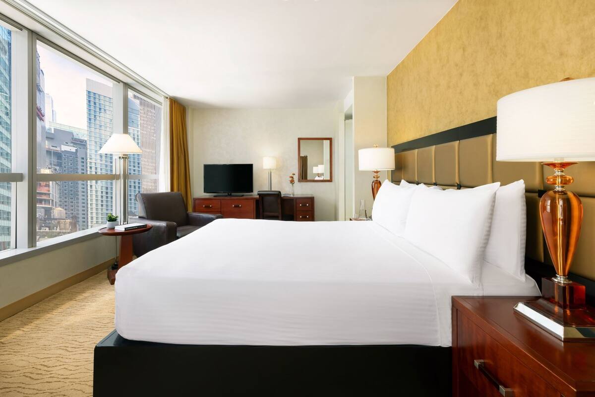 A hotel room with a large bed, a desk, a chair, a television, and a view of the cityscape.