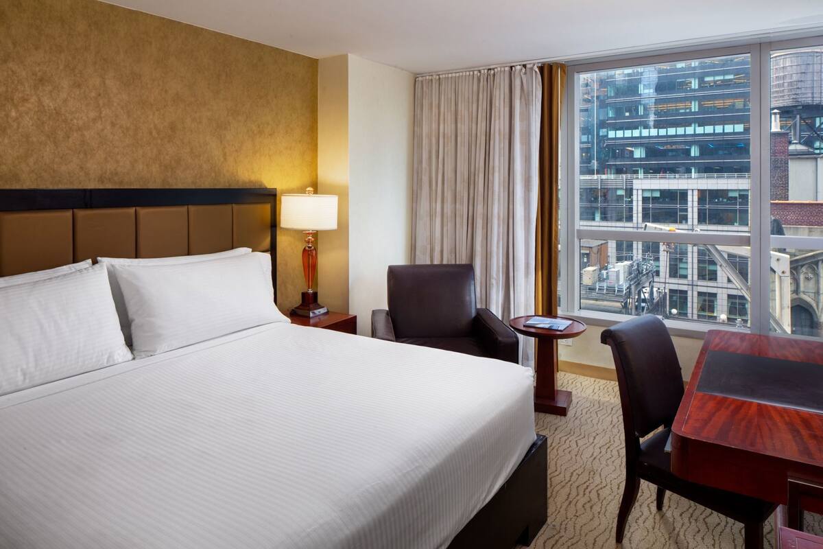 A hotel room with a large bed, a desk, two chairs, a lamp, and a view of a cityscape through a large window.