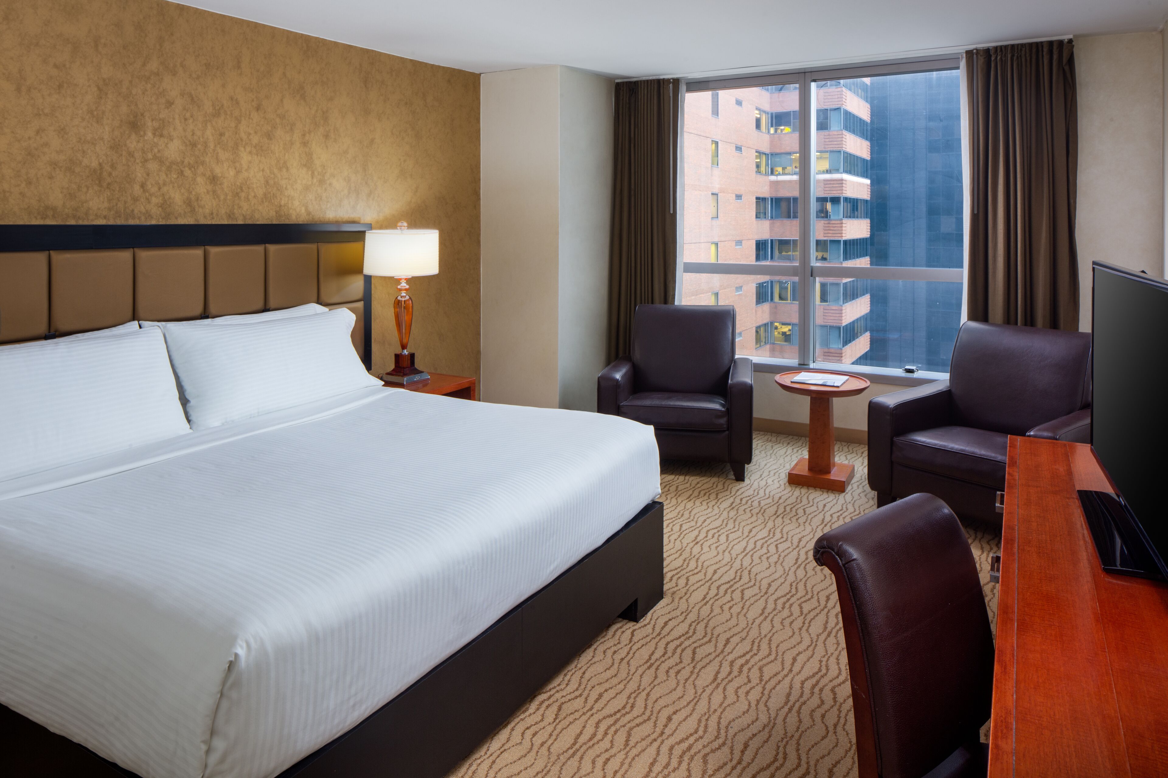 A hotel room with a large bed, two armchairs, a small table, a TV, and a window with a city view.