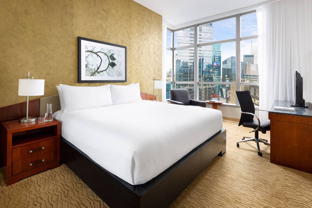 A hotel room with a large bed, a desk, a chair, a nightstand, a lamp, and a large window with a city view.