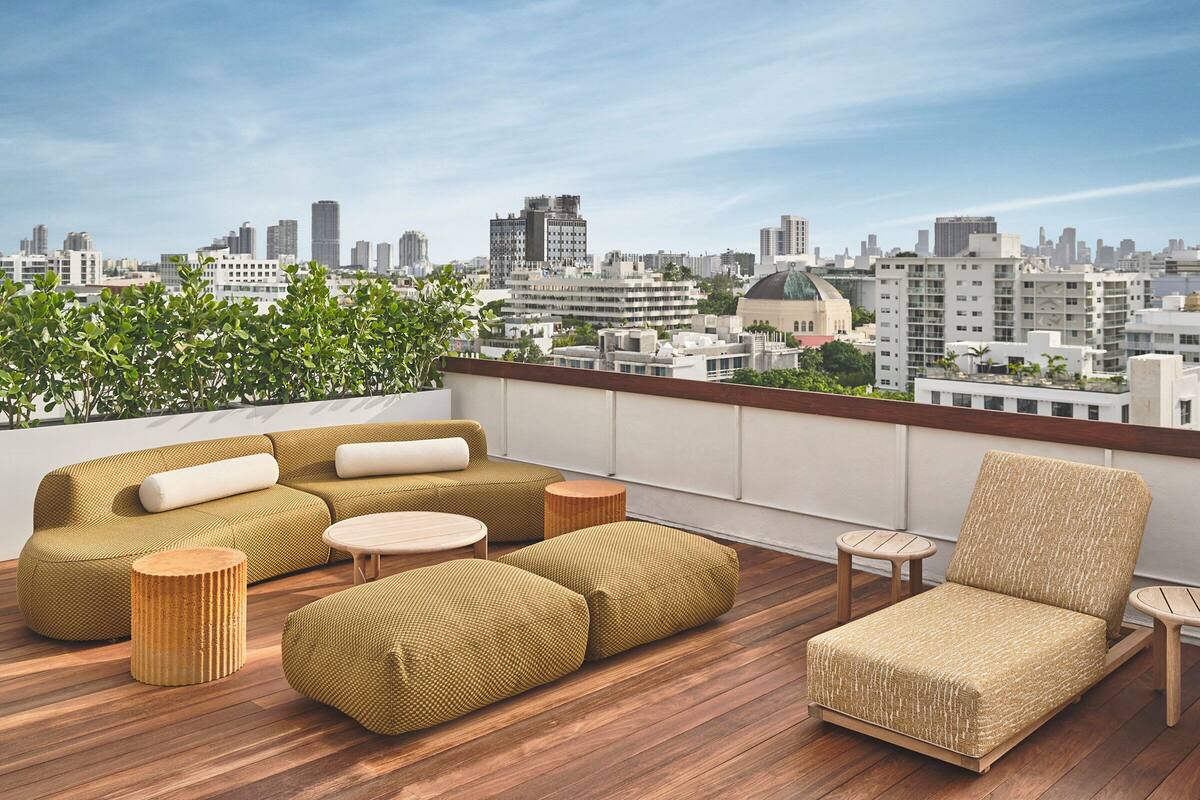 A rooftop terrace with a sofa set, ottomans, and a chair, overlooking a cityscape.