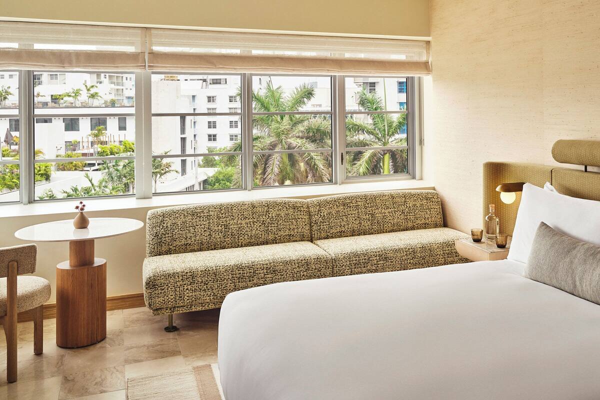 A hotel room with a large bed, a sofa, a round table, and a view of palm trees and buildings through large windows.