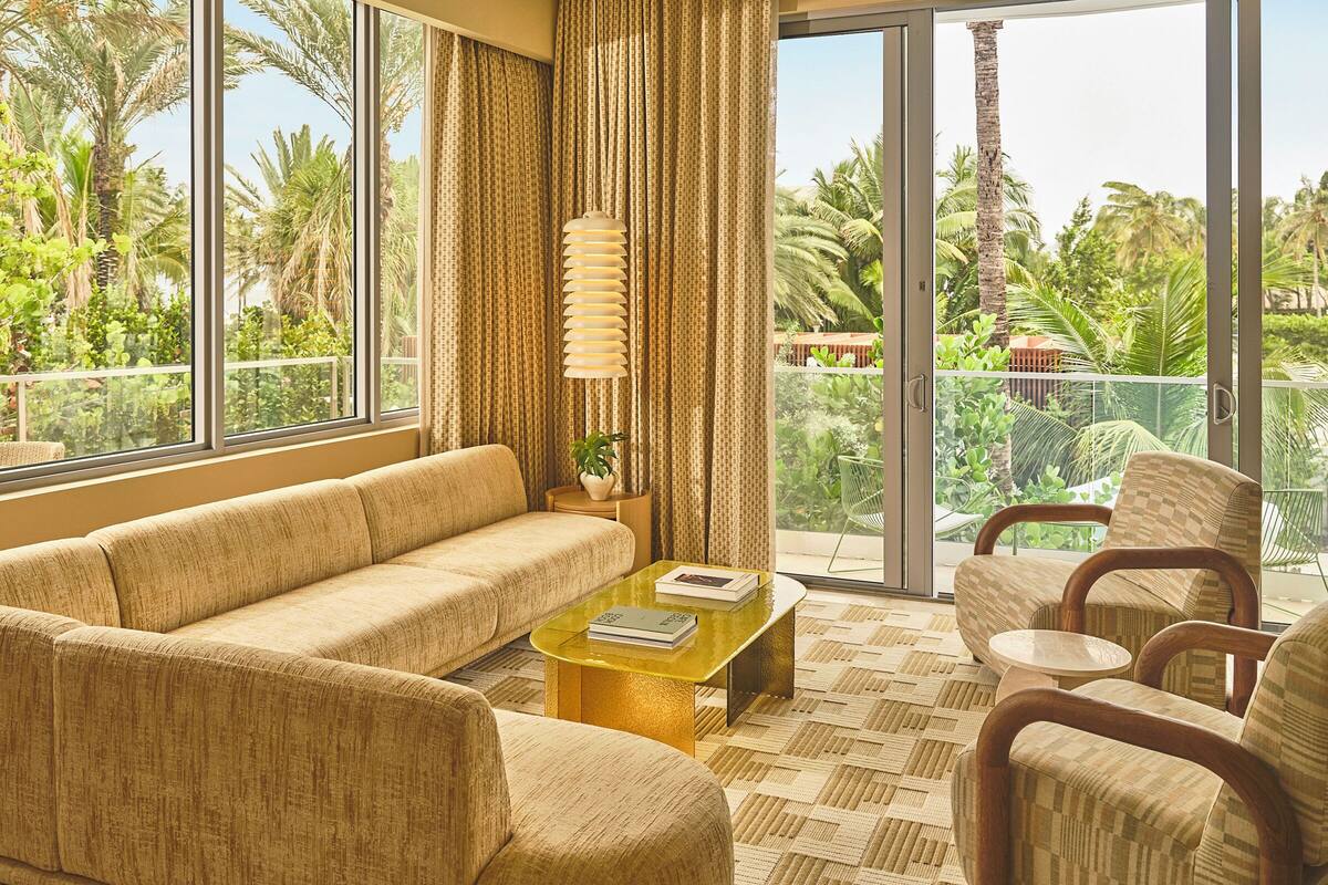 A spacious living room with a sofa, armchairs, and a coffee table. Large windows offer a view of lush greenery and palm trees.