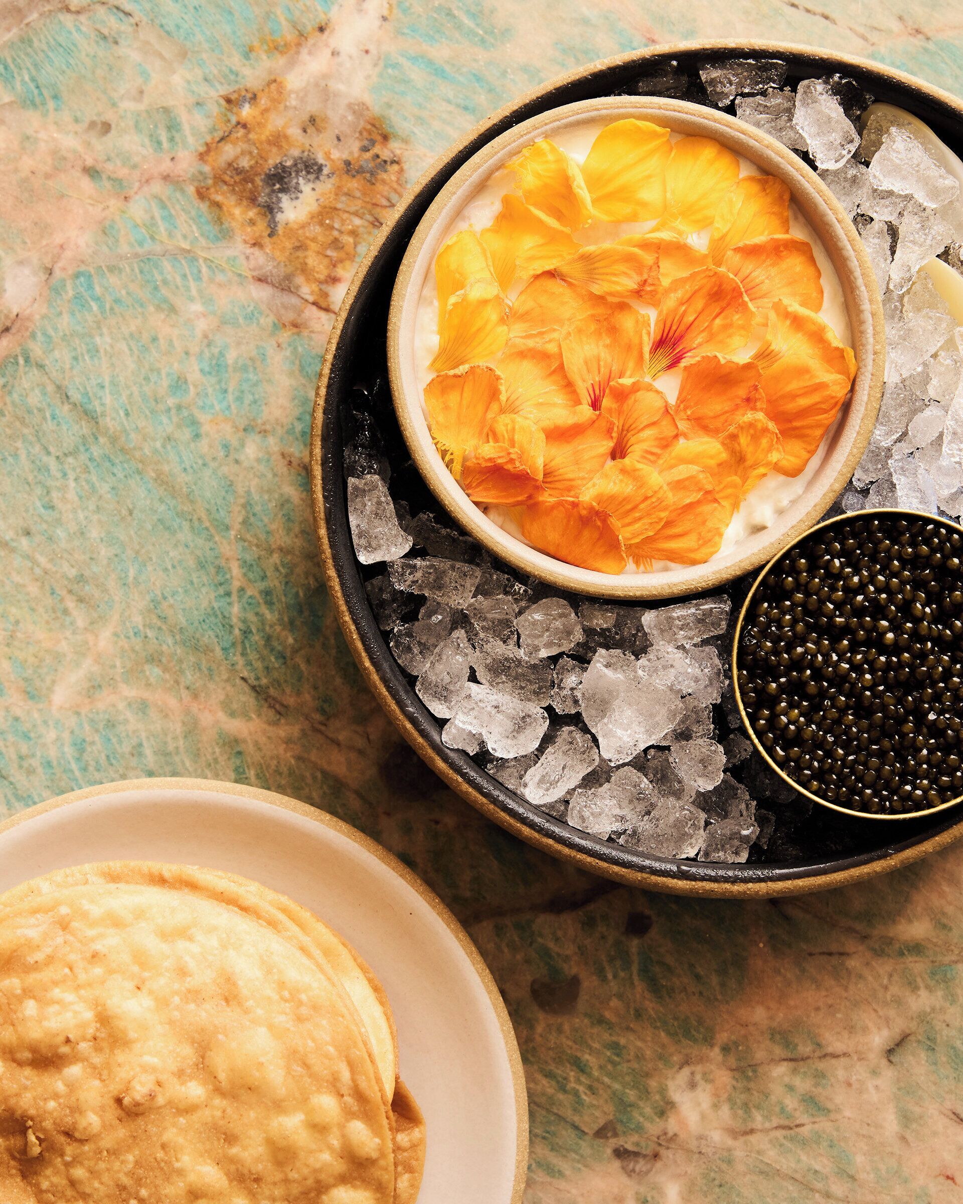 Un bol de pétales de fleurs d’oranger sur de la glace, un bol de caviar noir et une assiette de nourriture sur une surface texturée.