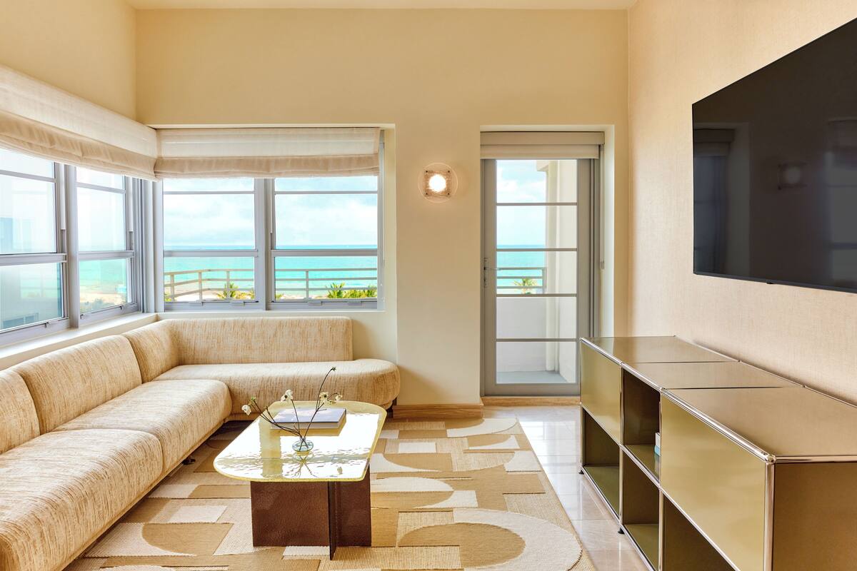 A modern living room with a large sofa, a coffee table, and a TV. There are large windows with a view of the ocean.