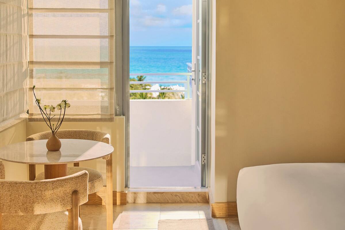 A room with a round table, a vase with flowers, and a view of the ocean through an open door.