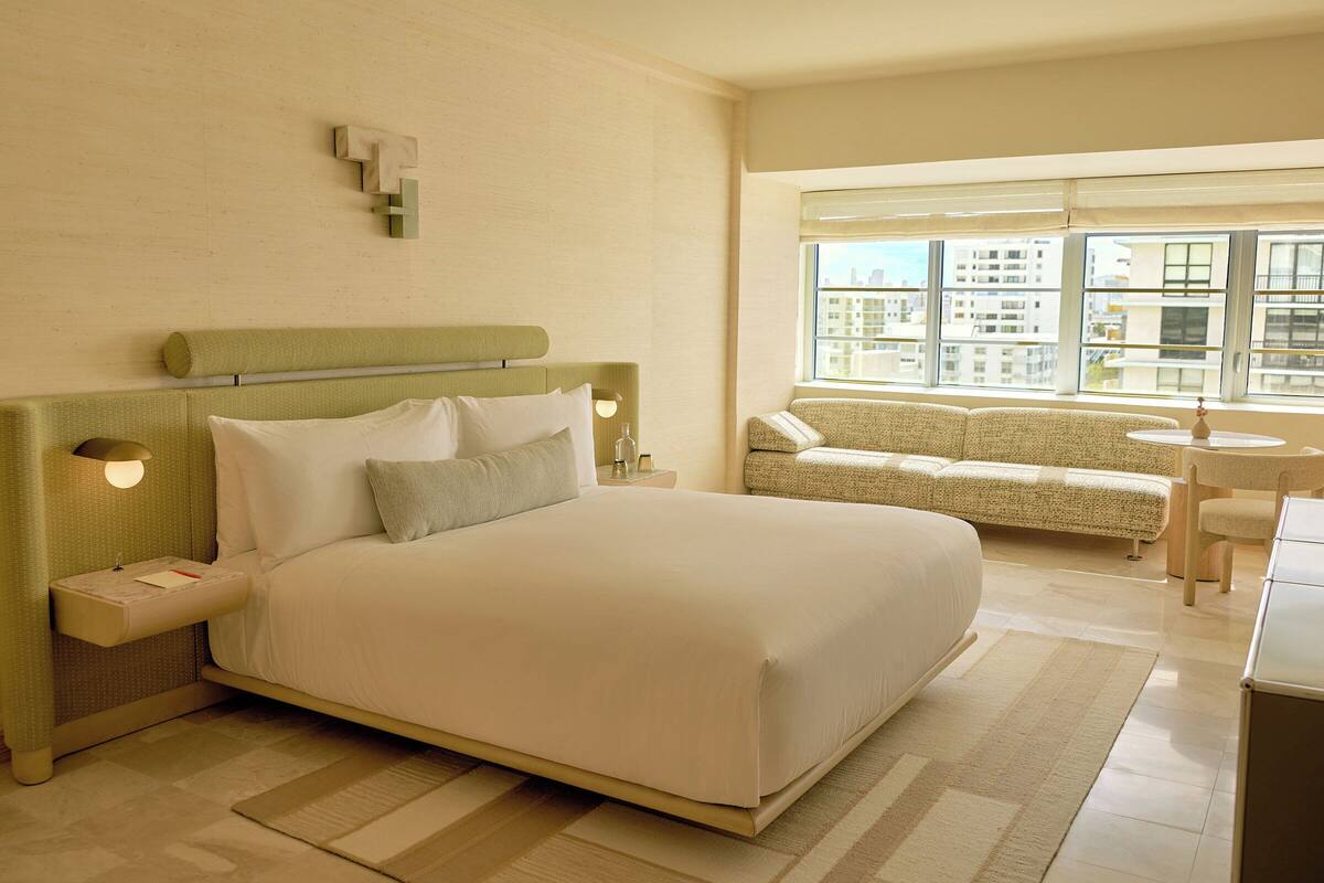 A hotel room with a large bed, a sofa, a small table, and a window with a city view.