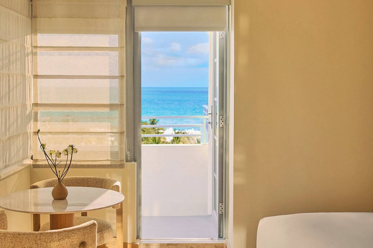 A modern hotel room with a large bed, a dining area with a table and chairs, a view of the ocean through a sliding door, and a small vase with flowers on the table.