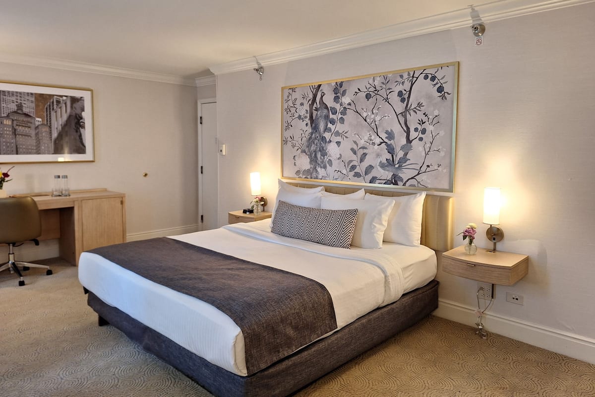 A neatly arranged hotel room with a bed, bedside tables, a desk, and a large framed artwork on the wall.