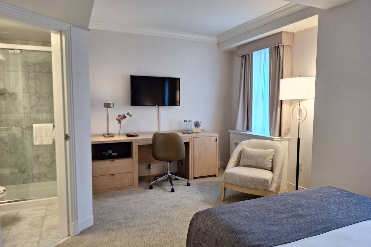 A hotel room with a bed, desk, chair, TV, and a bathroom with a shower.