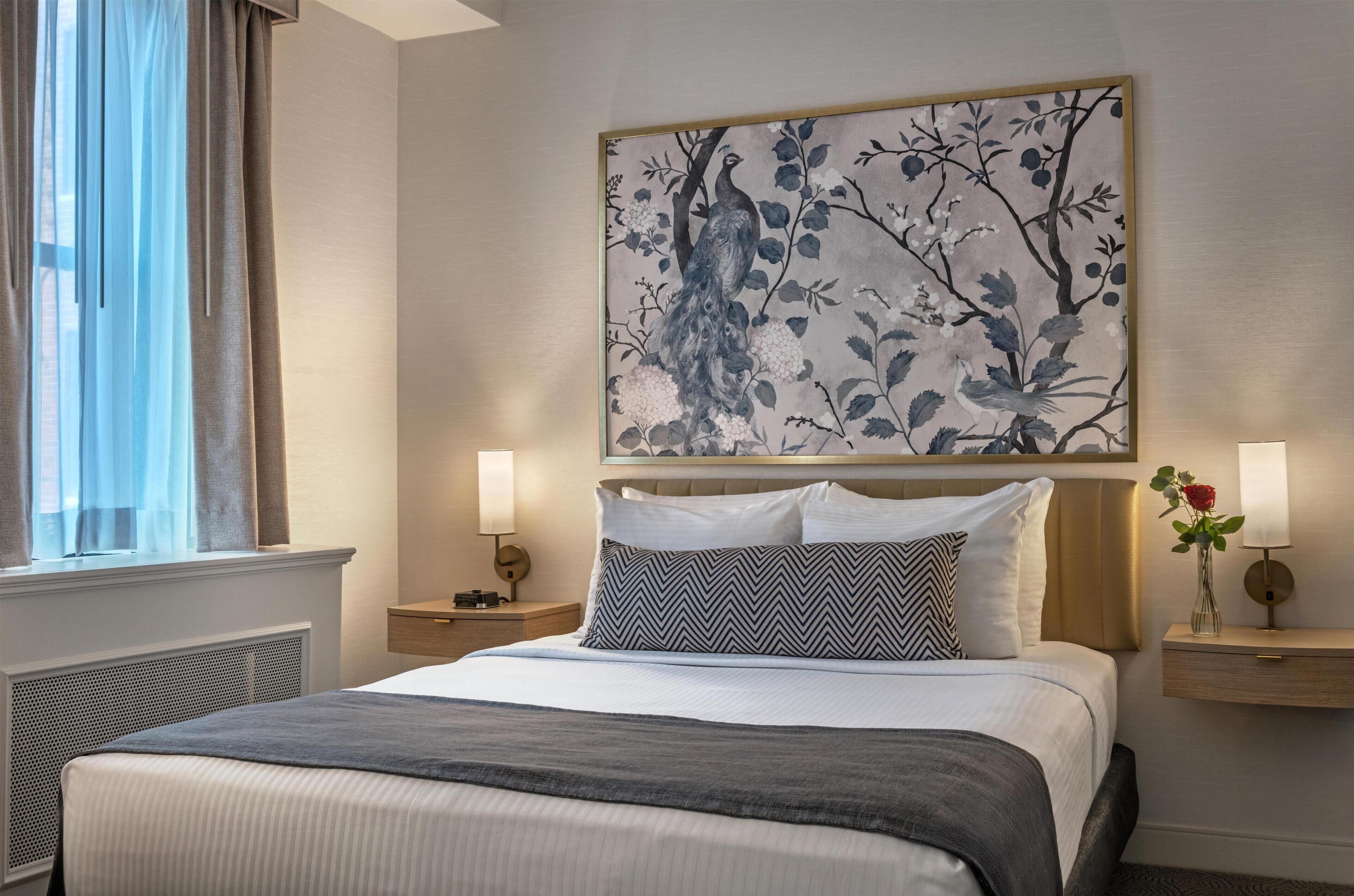 A hotel room with a bed, two bedside tables with lamps, a window with curtains, and a wall-mounted artwork.