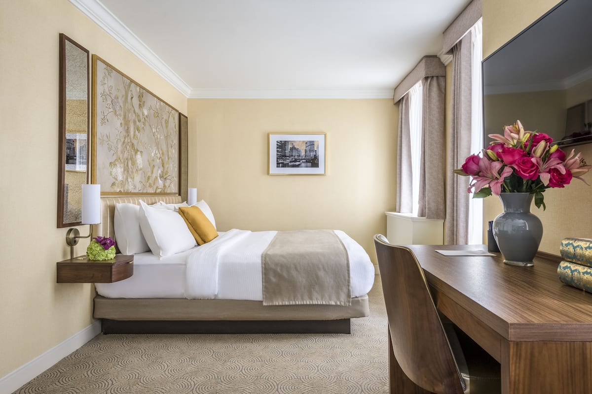 A hotel room with a bed, bedside table, lamp, and a vase with flowers.