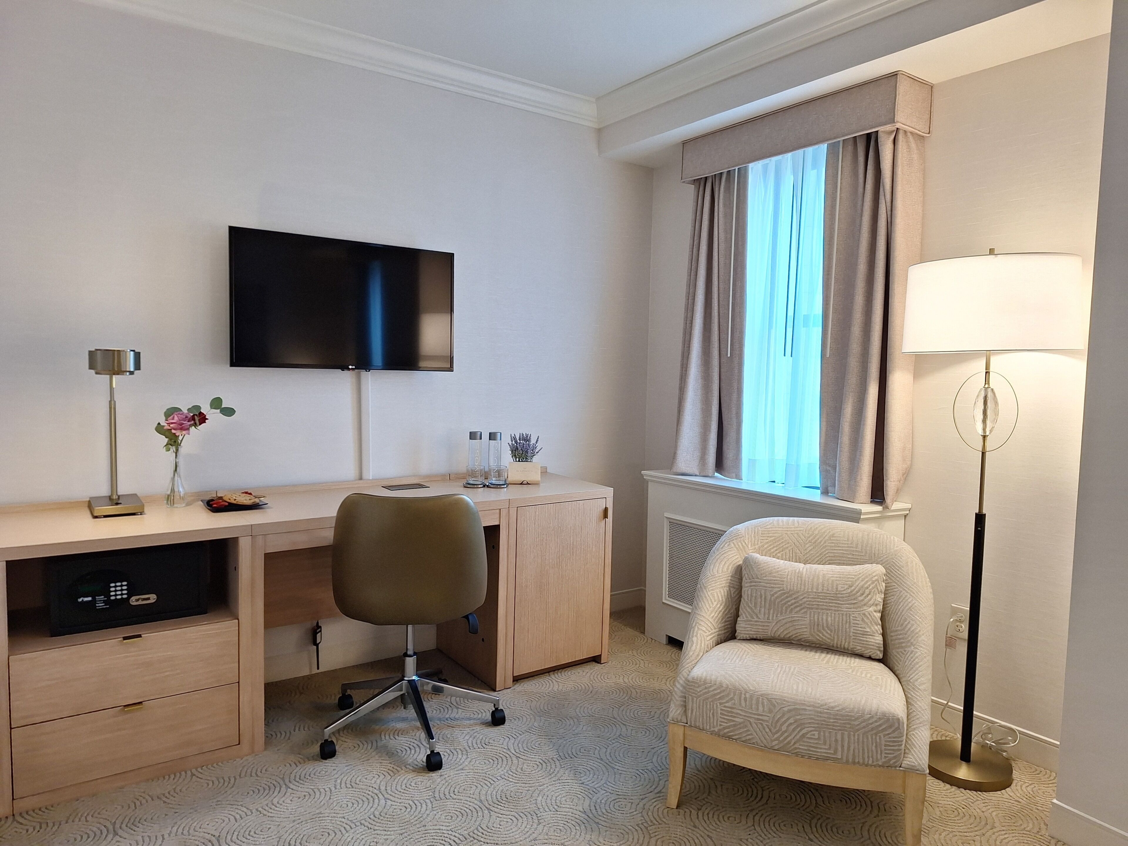 A hotel room with a desk, chair, TV, lamp, and a window with curtains.