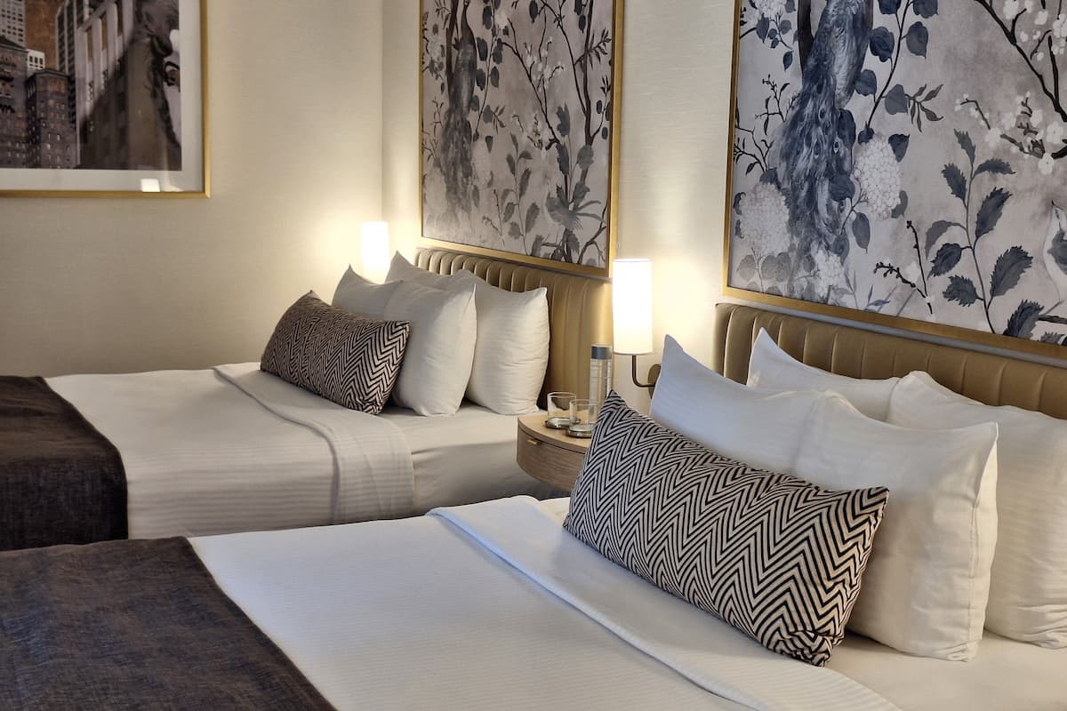 A hotel room with two beds, each with white linens and patterned pillows, a nightstand with a lamp, and framed artwork on the walls.