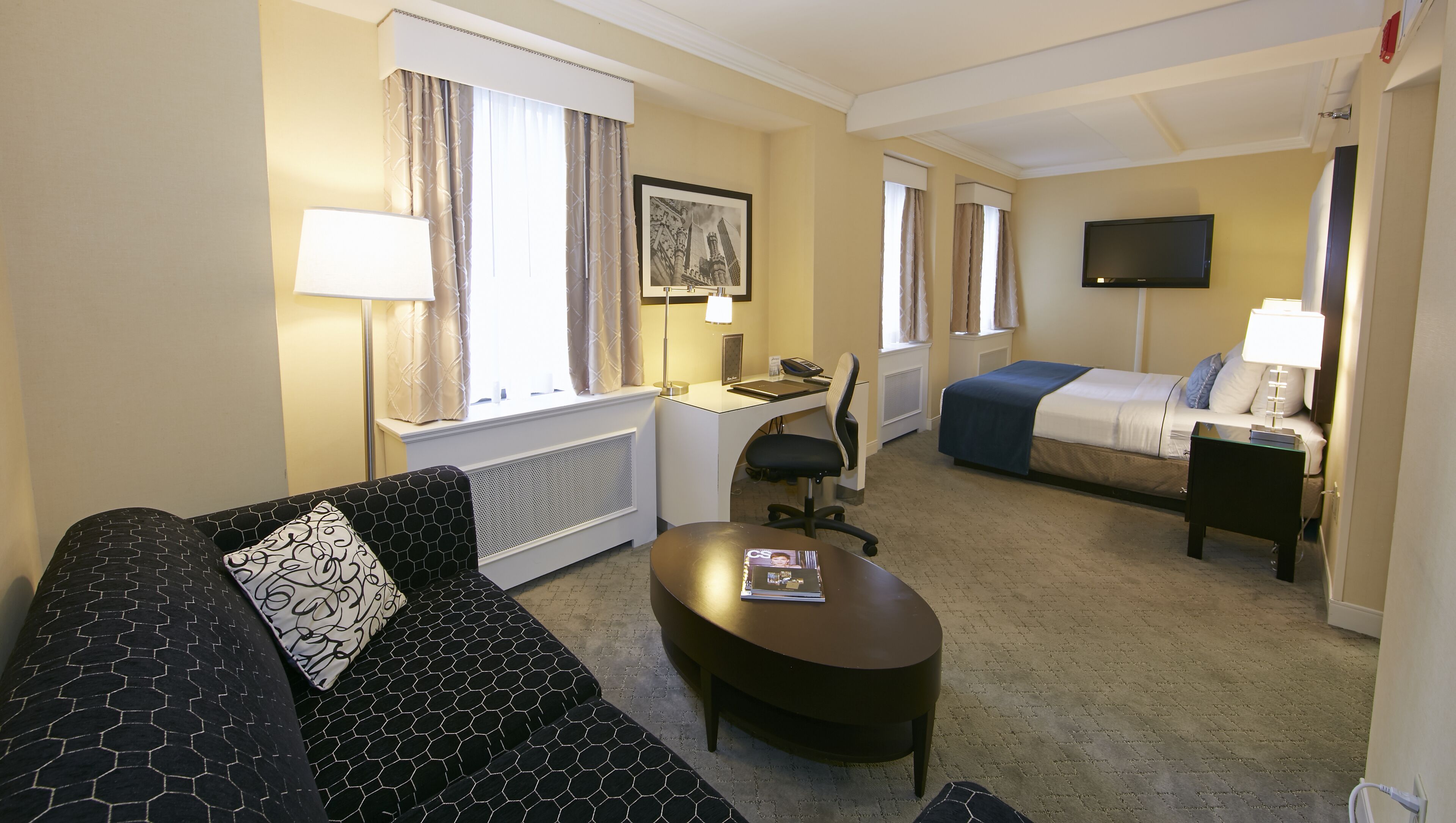 A hotel room with a sofa, a desk, a bed, a TV, and a window with curtains.