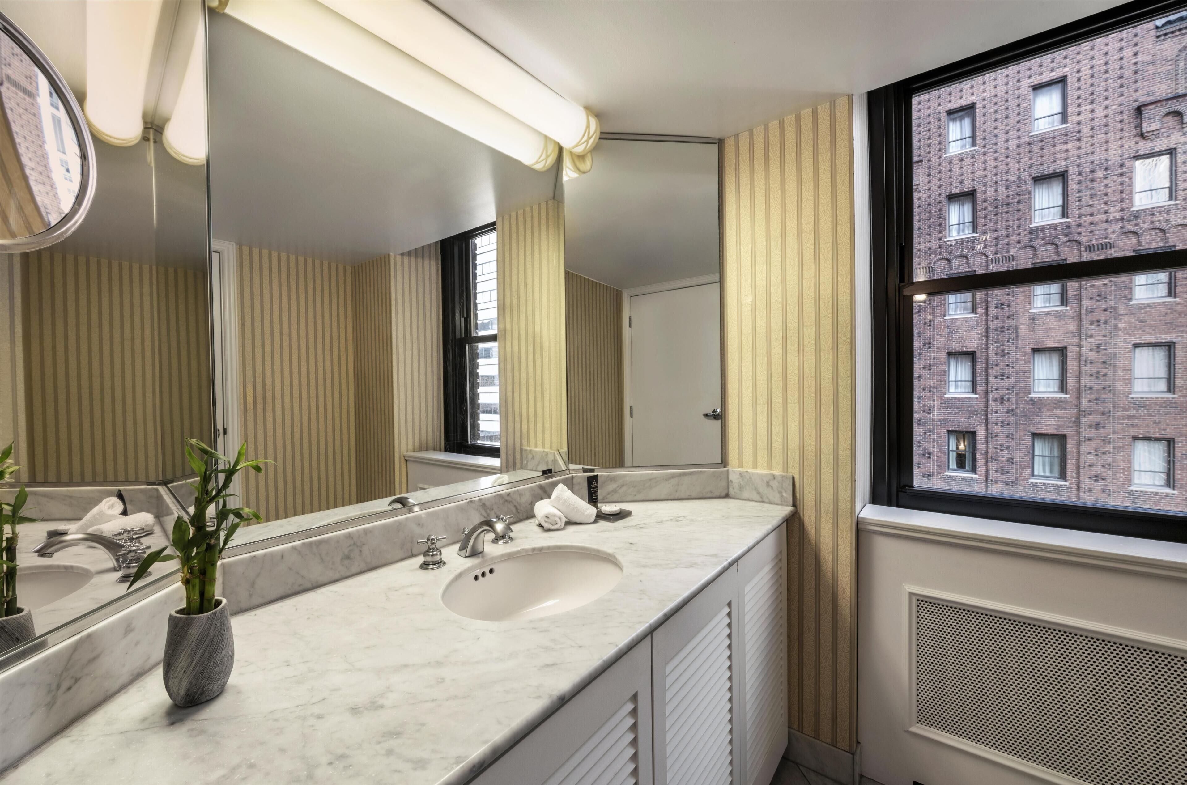A modern bathroom with a large mirror, two sinks, and a window with a view of a brick building.