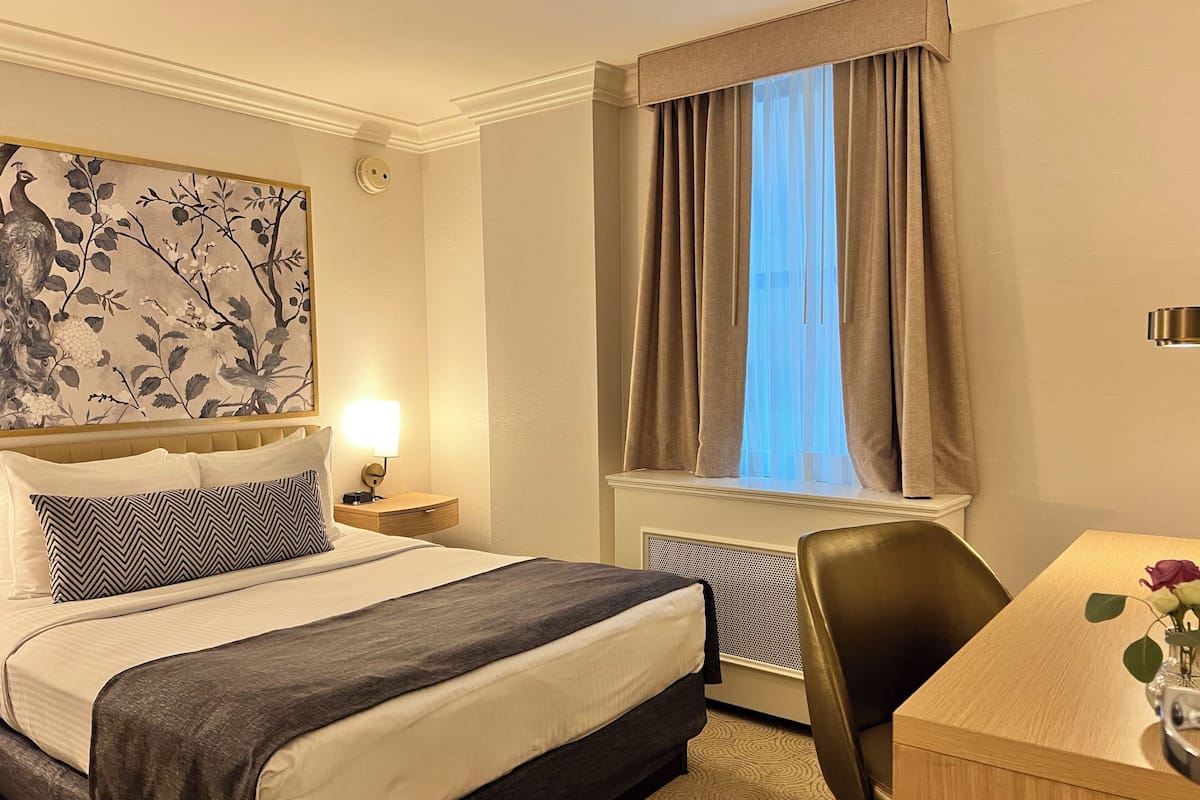 A hotel room with a bed, a desk, a chair, a radiator, a window with curtains, a wall-mounted lamp, and a floral artwork.